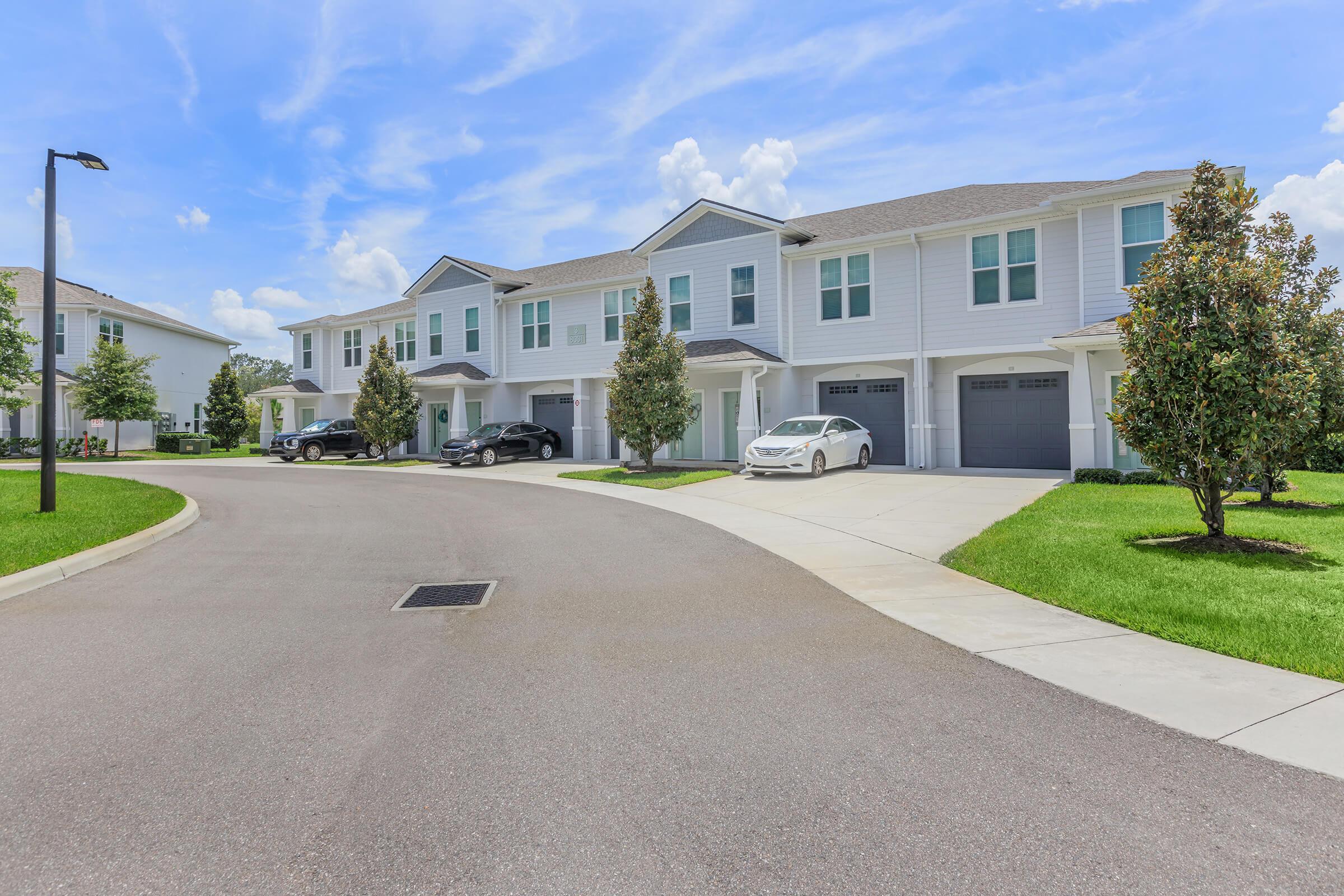 A picturesque residential street featuring modern townhouses. The well-maintained homes have grey and white exteriors, with landscaped lawns and young trees. Two cars are parked in driveways, and the sky is bright blue with fluffy clouds. The street curves gently, creating a serene neighborhood atmosphere.