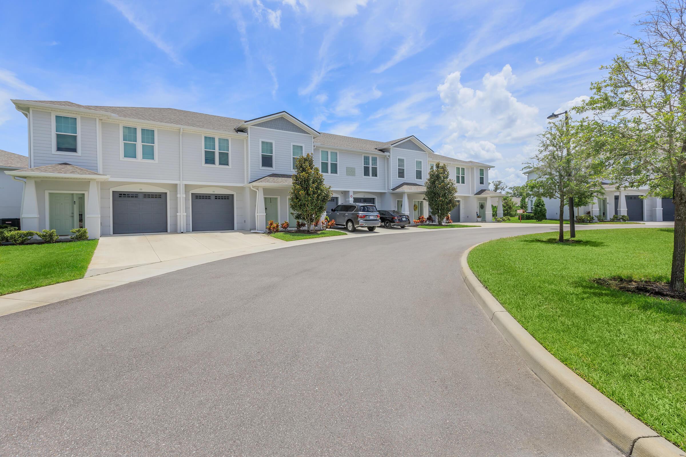 A peaceful residential street featuring modern townhouses with white exteriors and garages. The scene includes well-manicured lawns, neatly trimmed trees, and bright blue skies with fluffy clouds, creating a welcoming suburban atmosphere.