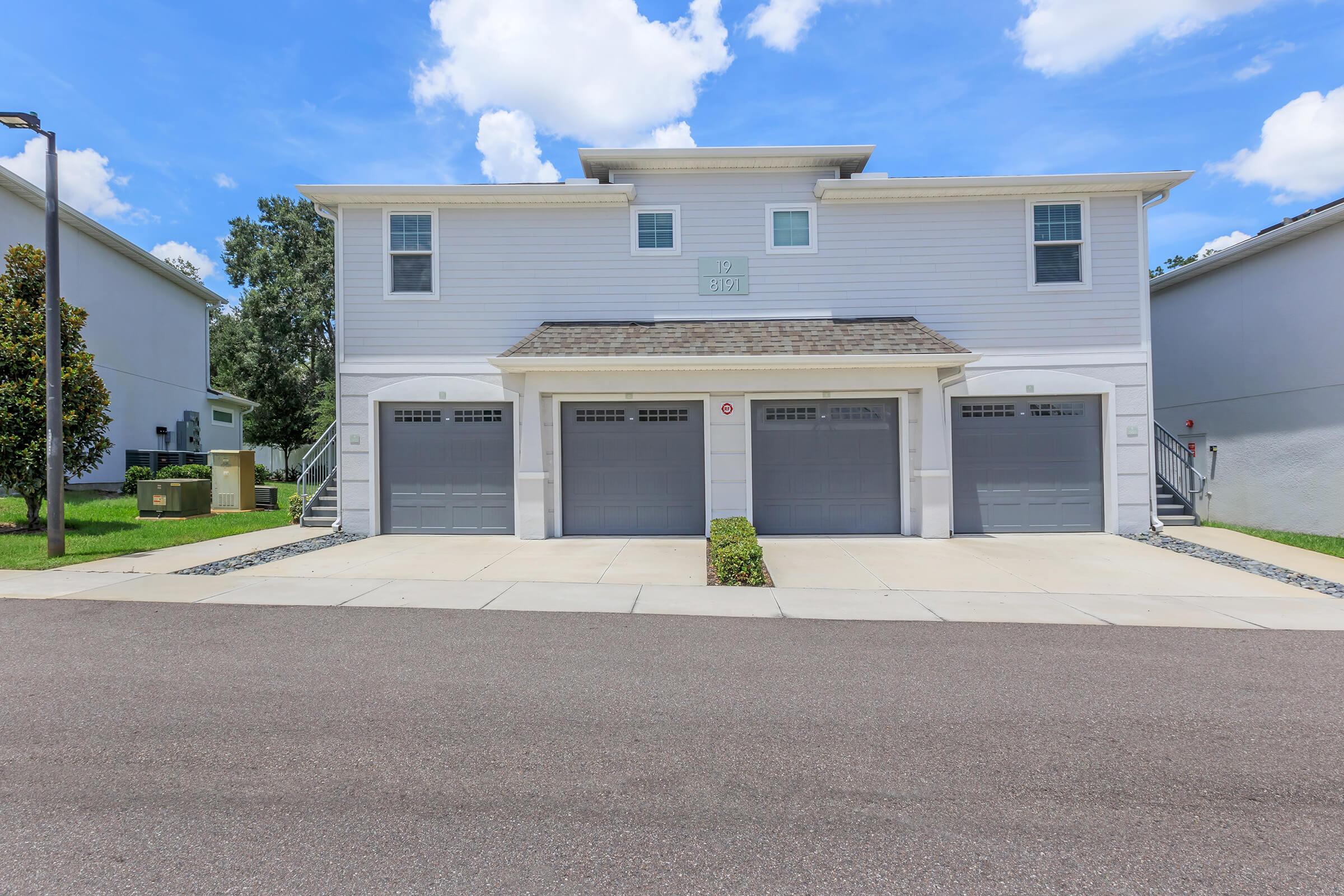 A two-story residential building with a light gray exterior, featuring three garage doors and a set of stairs on the left side. The street is paved, with greenery along the edges, and blue skies with scattered clouds above.