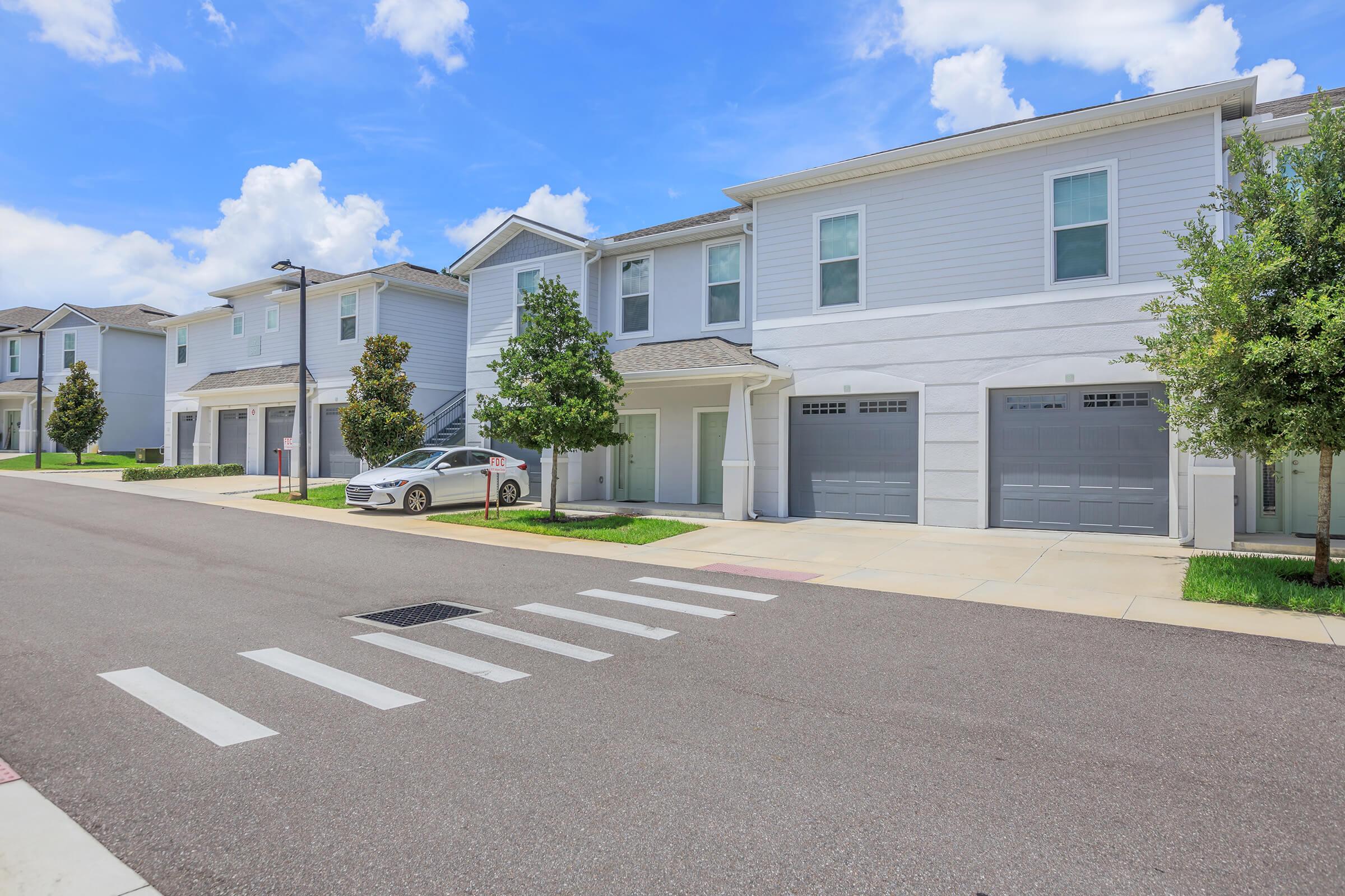 A quiet suburban street lined with modern townhouses. The buildings feature light gray exteriors with garages, well-maintained lawns, and trees. A clear blue sky with fluffy white clouds complements the peaceful atmosphere. A car is parked in the driveway, and a crosswalk is visible in the foreground.