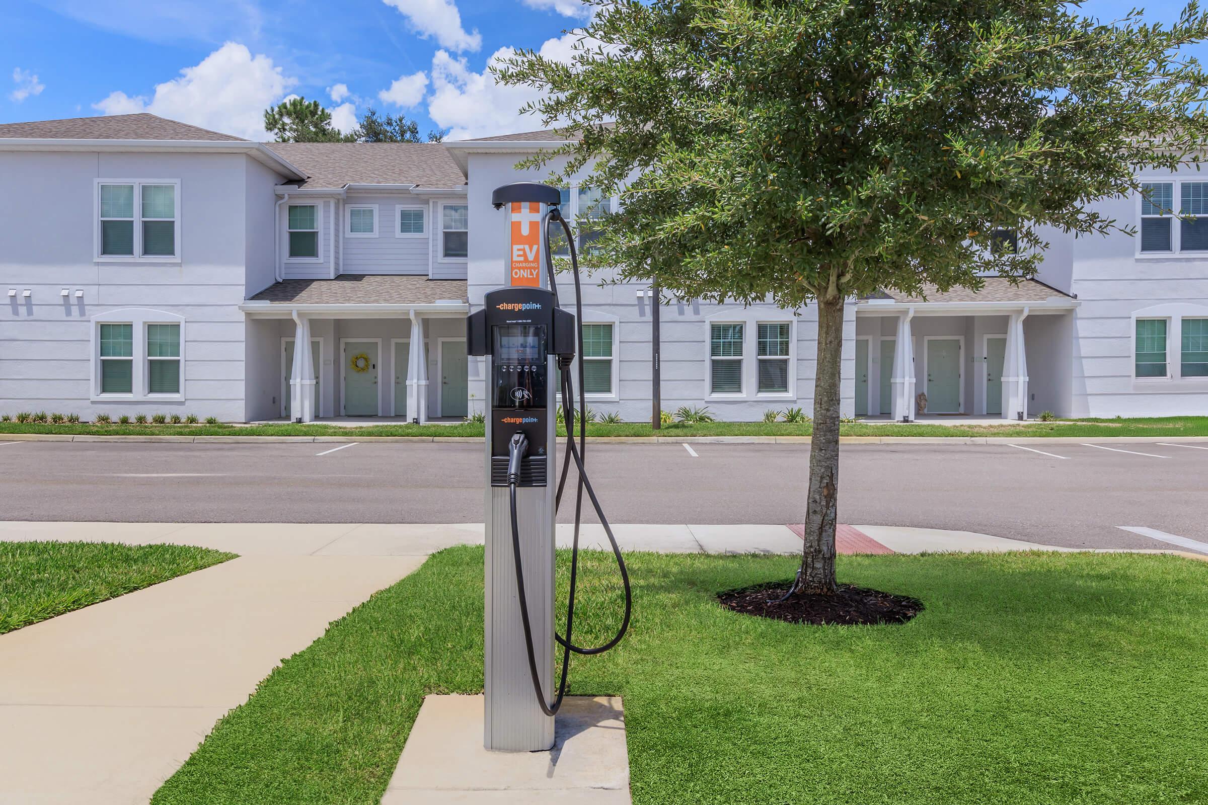 Electric vehicle charging station located on a sidewalk in front of a residential building. The station features a sleek design with a charger and two cables ready for use. Surrounding the station are well-maintained lawns and parking areas, with houses visible in the background under a clear blue sky.