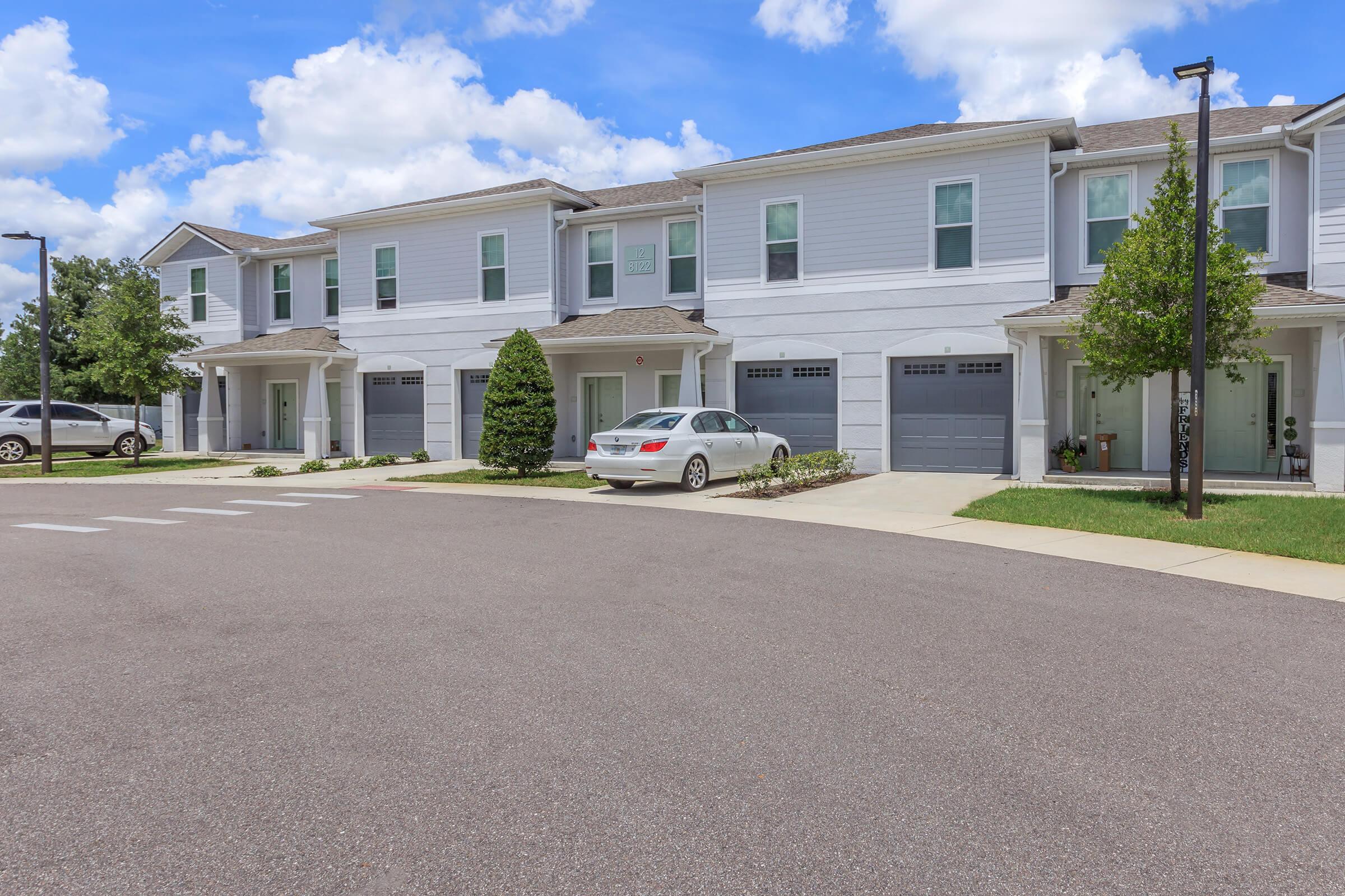 A row of modern two-story townhouses with garages, featuring light gray exteriors and manicured lawns. The driveway has a white car parked in front. The sky is partly cloudy, creating a bright atmosphere.