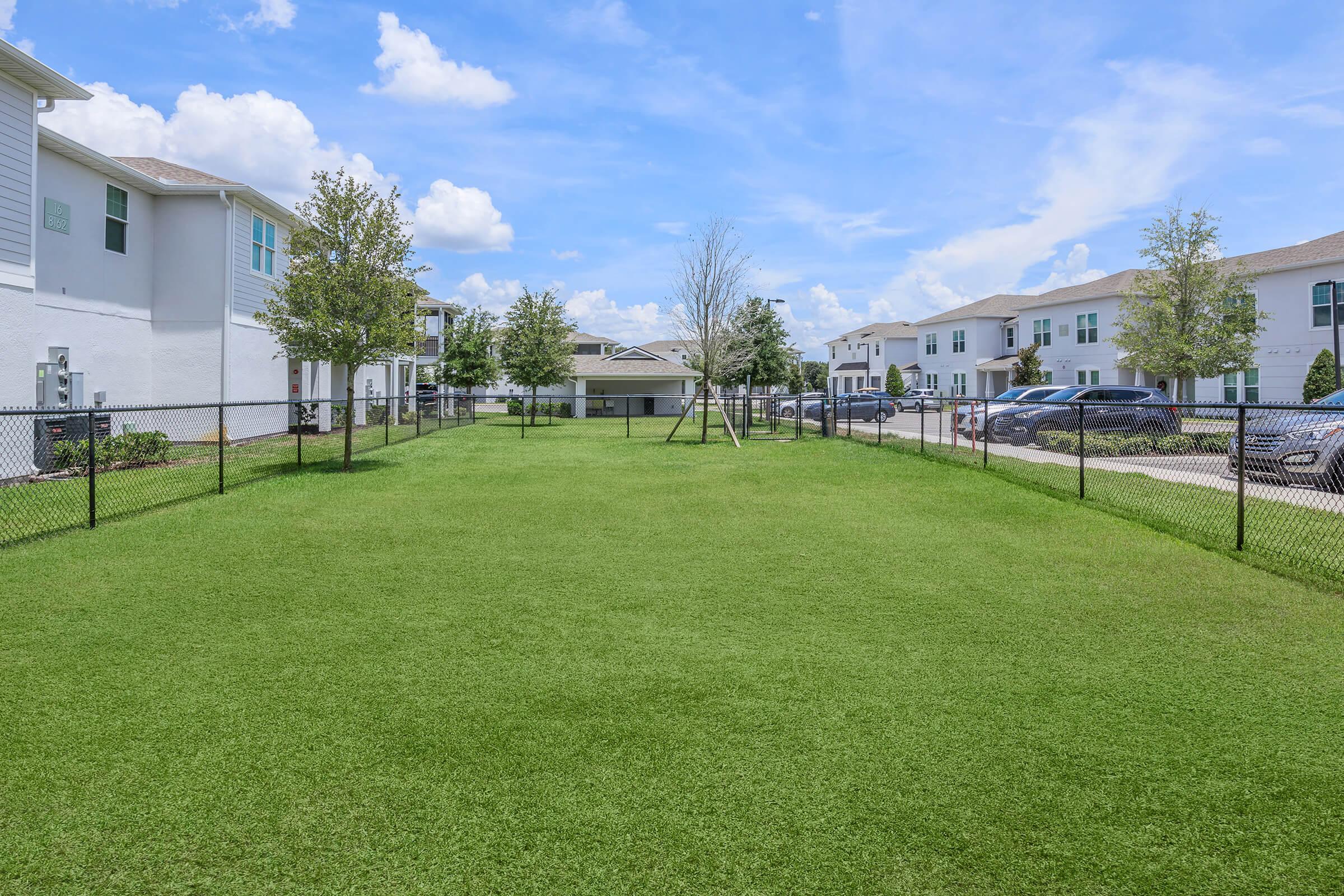 A well-manicured green lawn enclosed by a black fence, surrounded by residential buildings. The sky is bright with a few fluffy clouds, and there are young trees planted along the edges. Parking spaces are visible in the background along with nearby buildings.