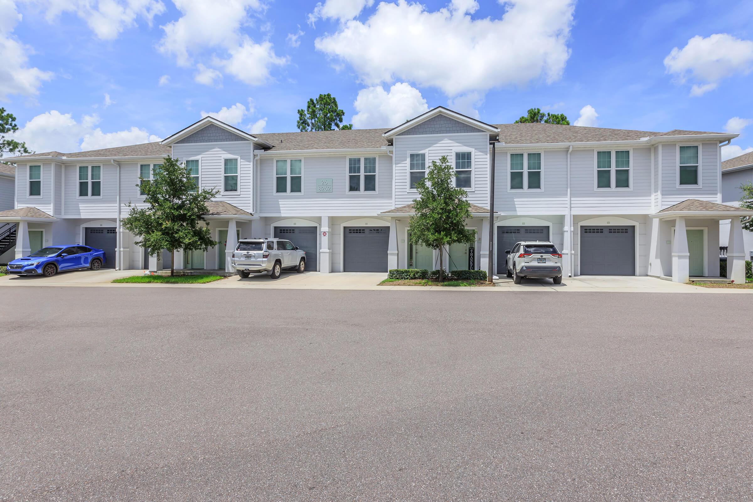 A row of modern, two-story townhouses with light blue siding and gray roofs. The buildings feature garages and are surrounded by well-maintained landscaping. Several cars are parked in front, and the sky is partly cloudy with vibrant blue hues.