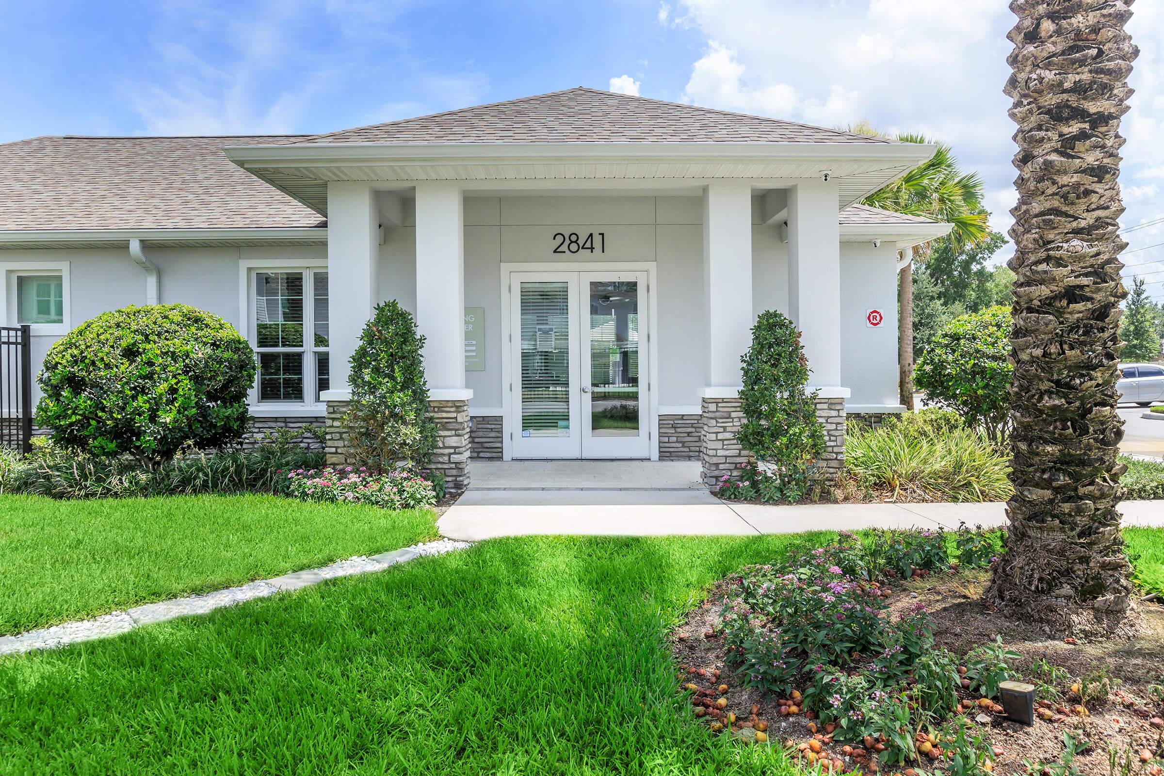 Front entrance of a modern building featuring a light gray facade, stone accents, and large windows. The door is centered with the number "2841" above it. There are neatly trimmed shrubs and flower beds on either side, along with a palm tree, and a well-maintained lawn in bright green. Clear blue sky with a few clouds in the background.
