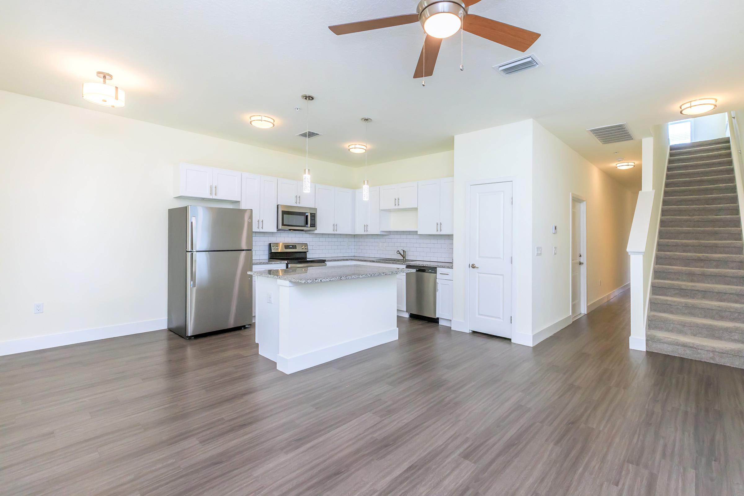 Modern open-concept kitchen and living area featuring stainless steel appliances, a granite countertop island, and a ceiling fan. Light-colored walls and wood-style flooring contribute to a bright, spacious feel. A staircase is visible in the background, leading to the upper level.