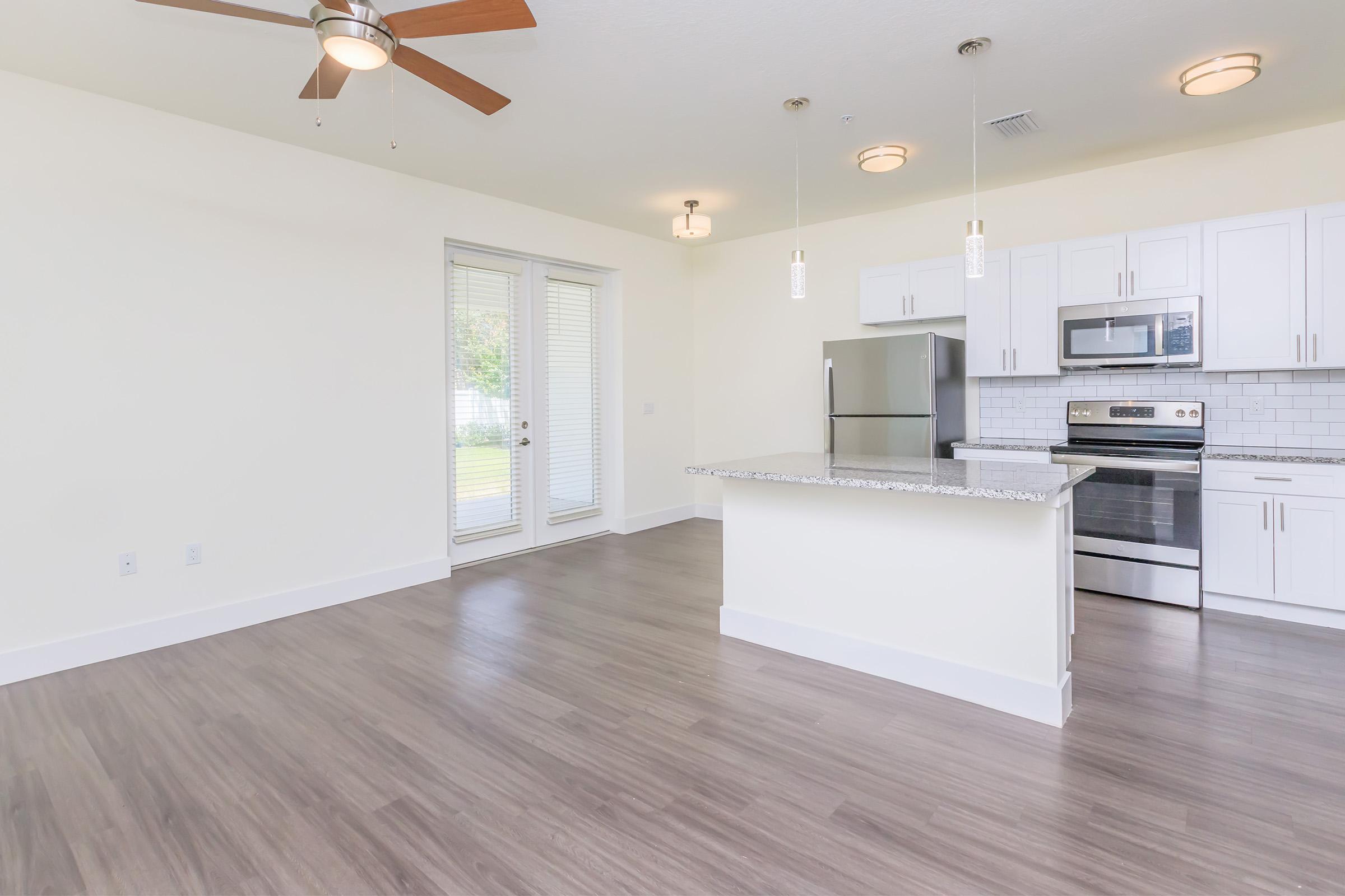 Bright and spacious modern kitchen and living area featuring stainless steel appliances, granite countertop, and light wood flooring. Large windows provide natural light, and a ceiling fan offers air circulation. White cabinetry complements the contemporary design.