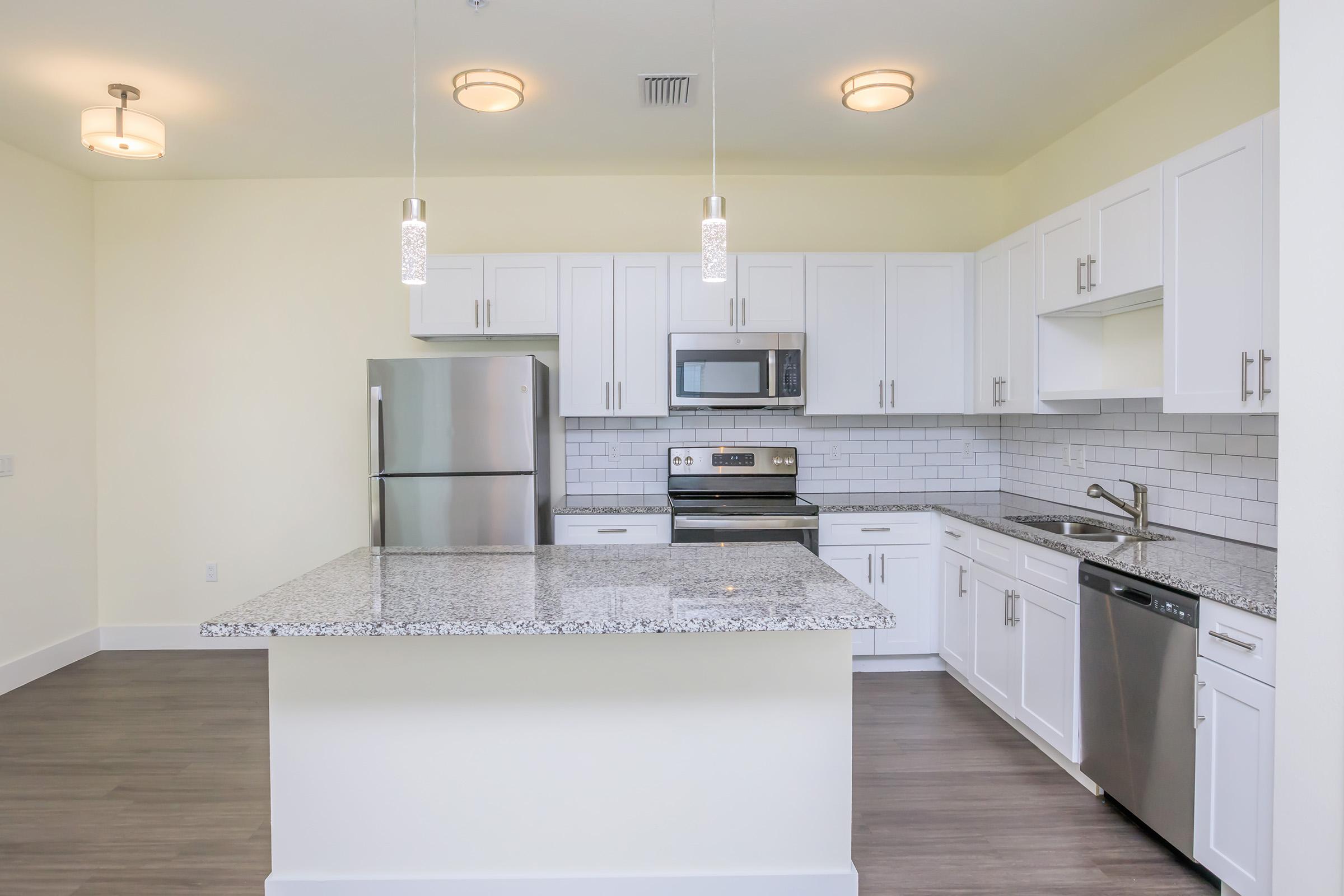 Modern kitchen featuring white cabinetry, stainless steel appliances, including a refrigerator and microwave, and a granite countertop with a central island. The backsplash is composed of white tiles, and the space is illuminated by pendant lights and natural light from the windows.