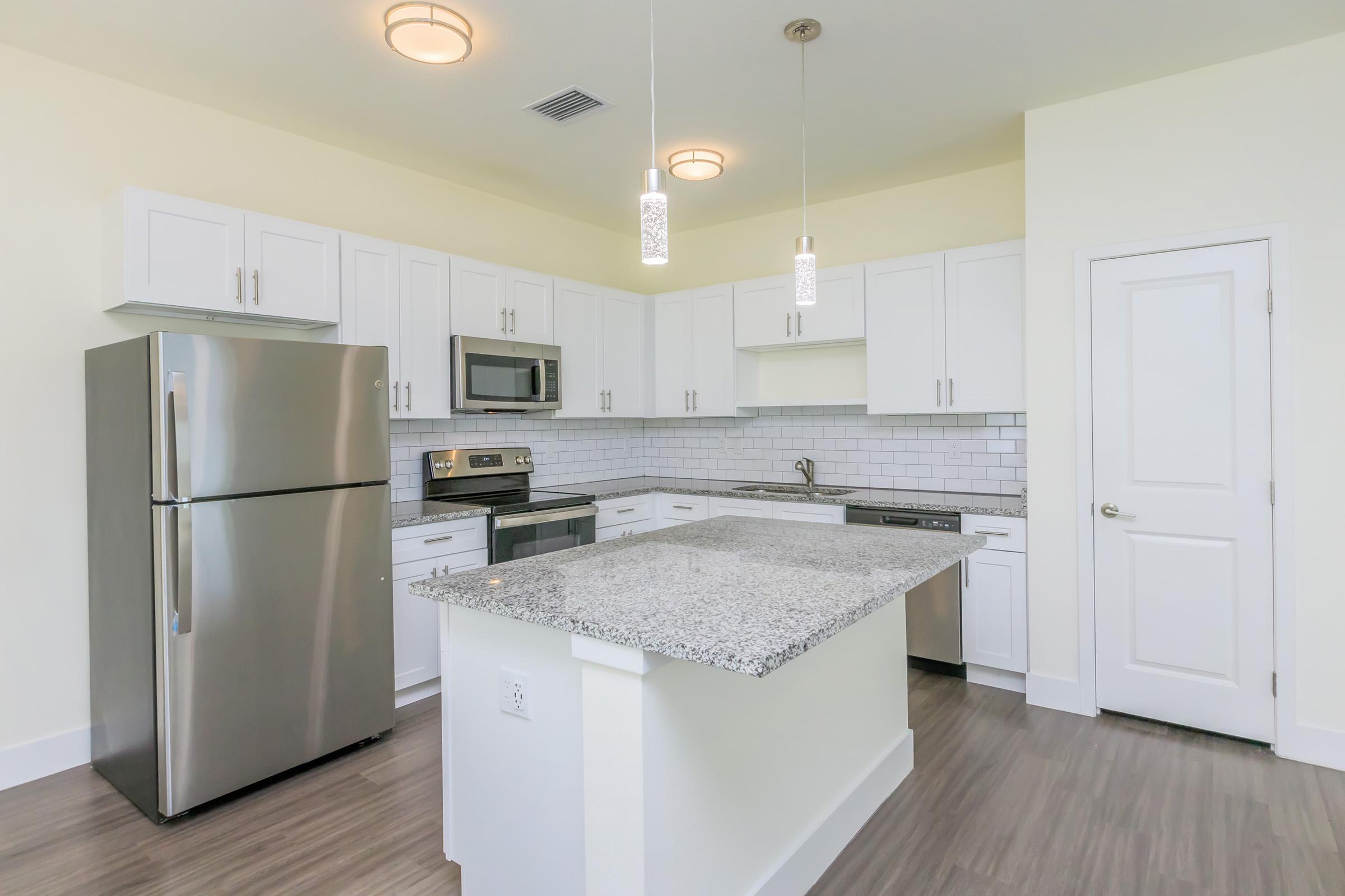 Modern kitchen featuring white cabinets, stainless steel appliances, and a granite island. The space includes a refrigerator, microwave, and a contemporary backsplash with pendant lighting above the island. The flooring is a sleek laminate, and the walls are painted in a light, neutral color.