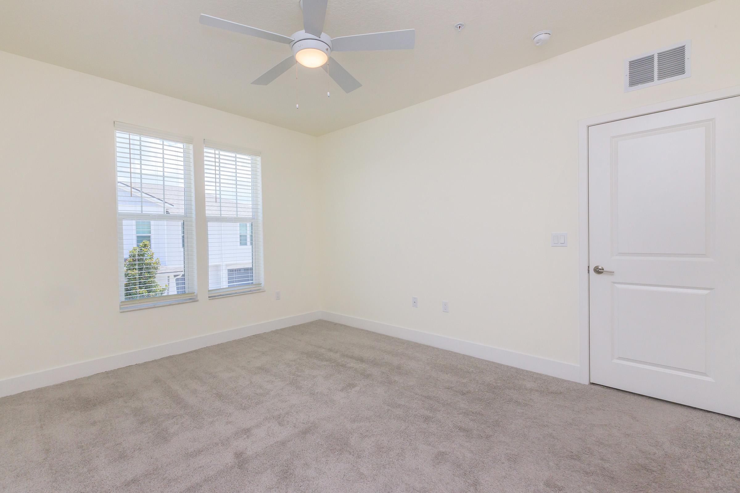 A well-lit empty room with beige carpet, a ceiling fan, and two windows with closed white blinds. The walls are painted in a light color, and there is a white door in the corner. The space is uncluttered, creating a minimalist atmosphere suitable for various uses.