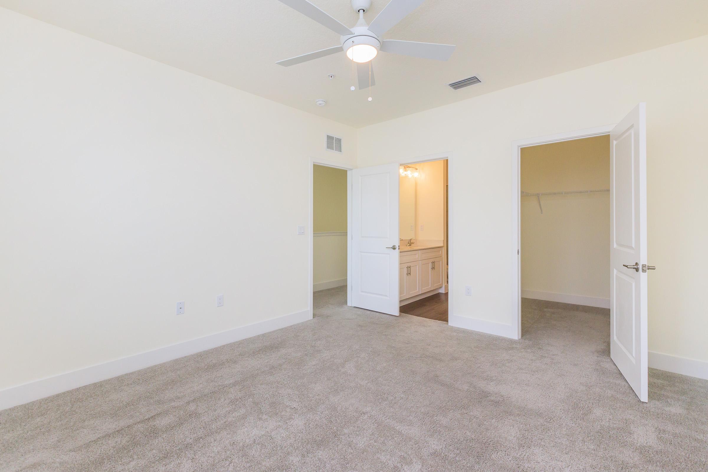 Empty bedroom featuring light-colored walls and carpeted flooring. A ceiling fan is visible, and two doors lead to a bathroom and closet, respectively. The overall ambiance is bright and spacious.