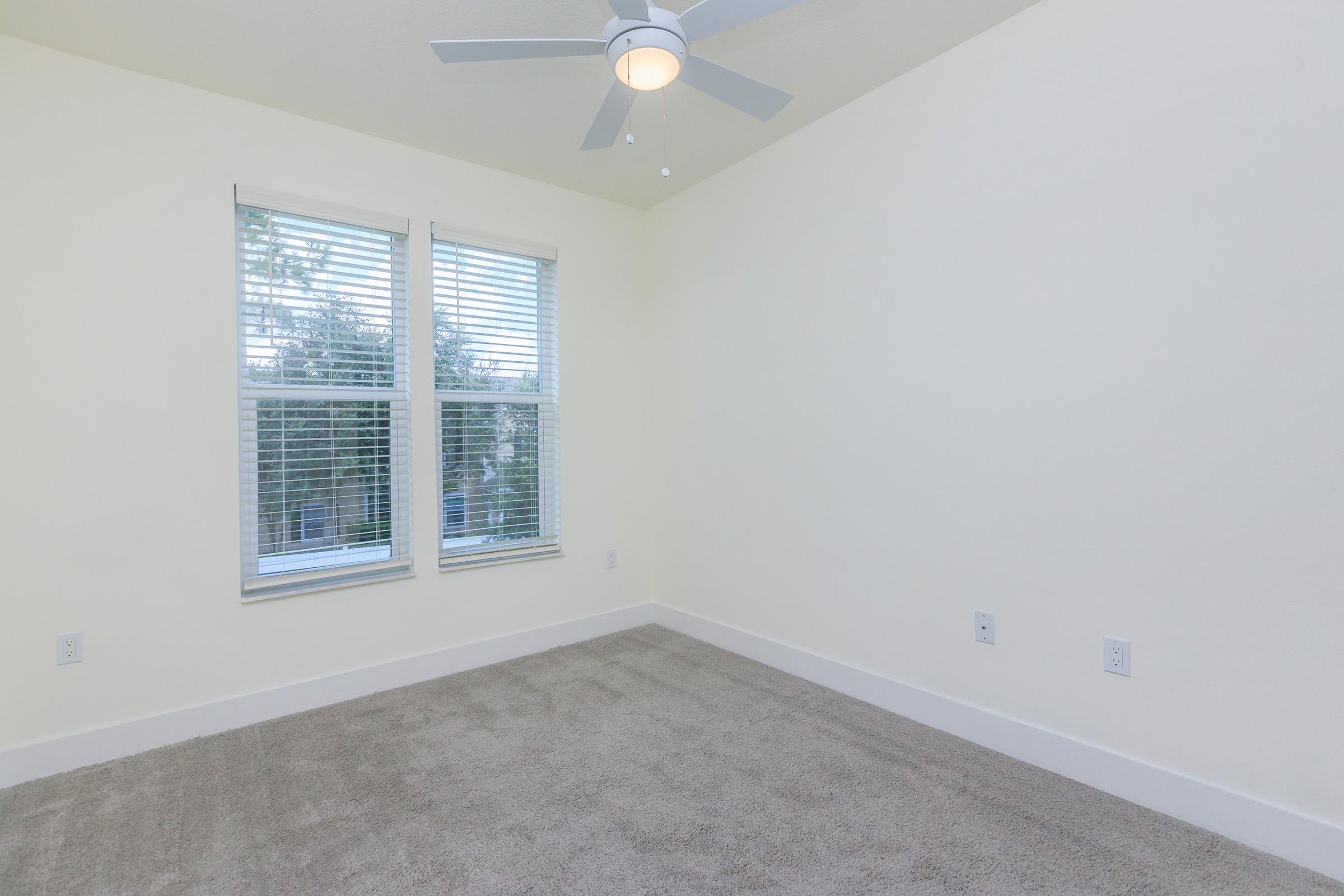 A well-lit empty room with light-colored walls and a ceiling fan. Two windows with blinds allow natural light to filter in, and the floor is covered with soft, gray carpeting. The room is uncluttered, creating a spacious and inviting atmosphere.