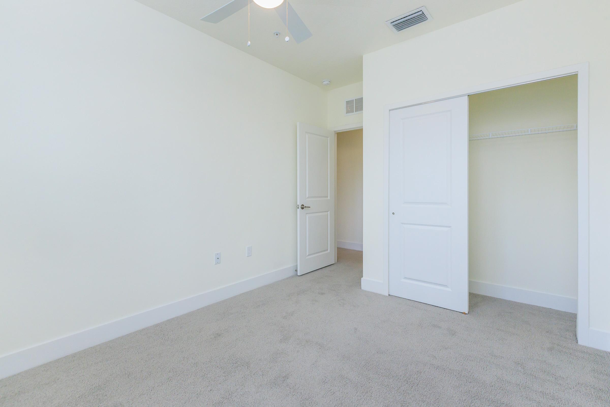 A light-colored room with a textured beige carpet, featuring a ceiling fan and a white door leading to a closet. The walls are a soft cream color, and the space is well-lit, creating an airy atmosphere. There are no furnishings, emphasizing the room's emptiness and potential for personalization.