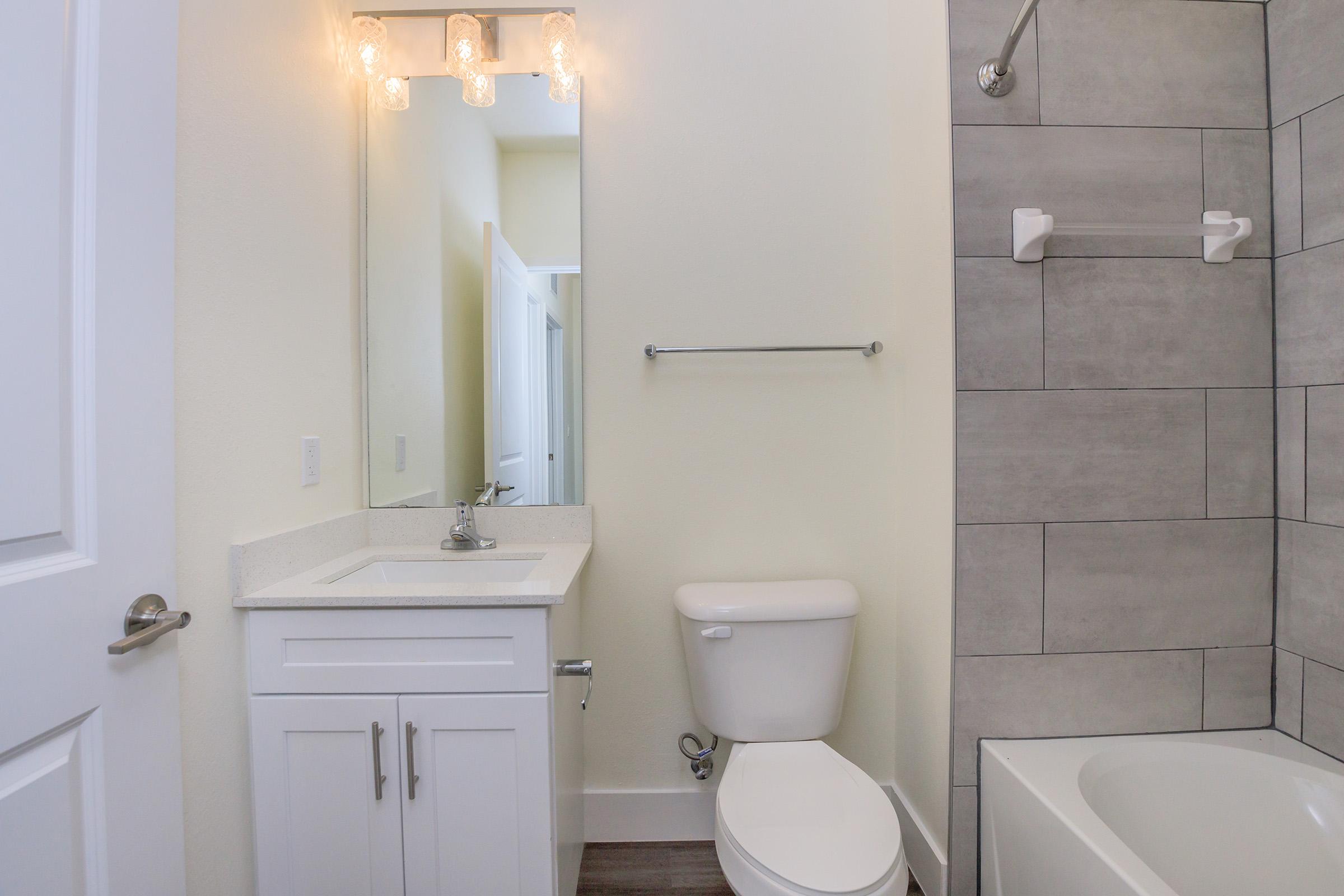 Modern bathroom featuring a white toilet, a sleek vanity with a sink, a rectangular mirror above the vanity, and a bathtub partially visible in the corner. Grey tiled walls add a contemporary touch, and the space is well-lit with stylish light fixtures above the mirror.