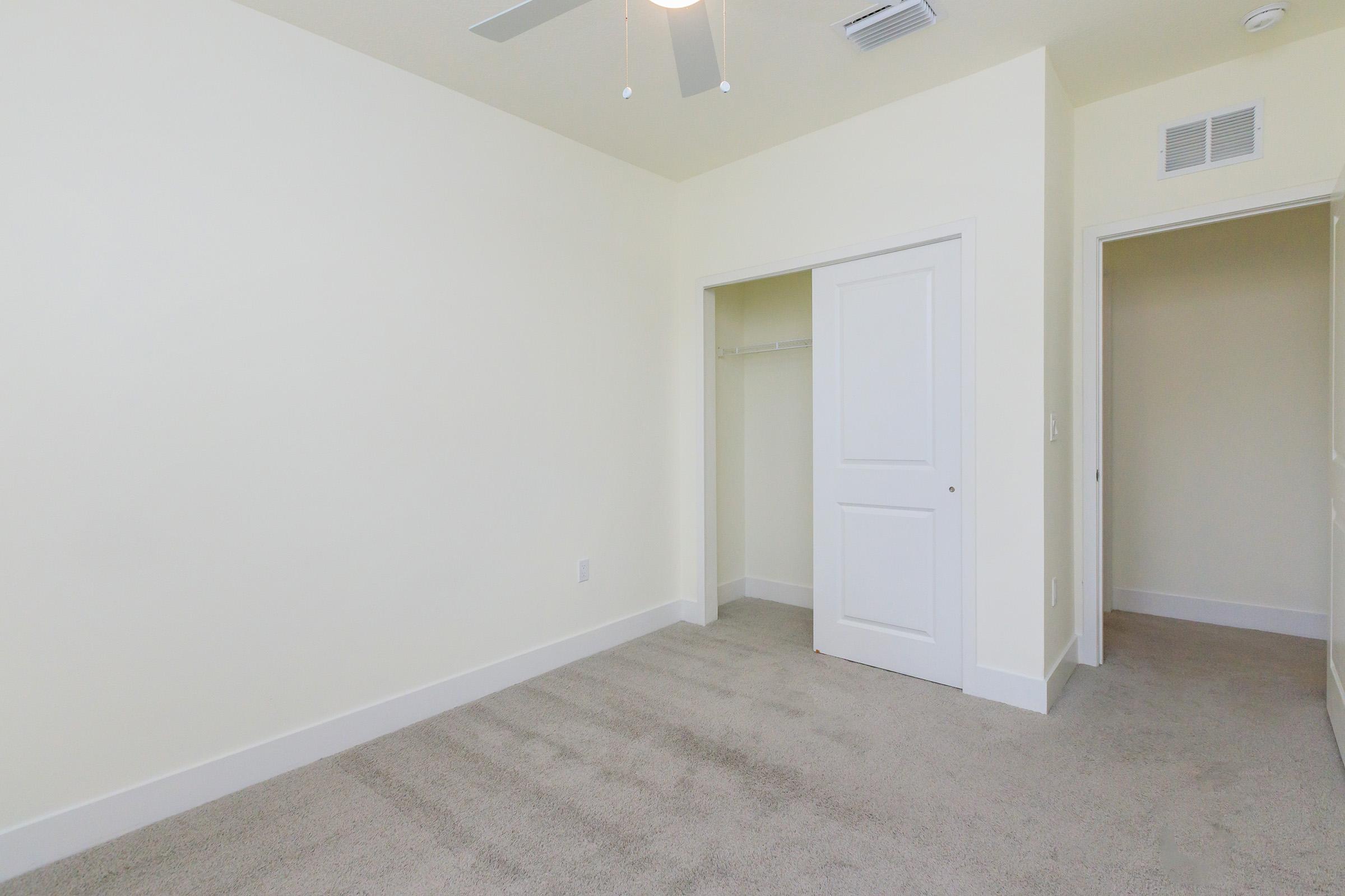 Empty room featuring light-colored walls, a ceiling fan, and a plush carpet. It includes a closet with a white door and another doorway leading to a hallway or another room. The overall design is clean and minimalist, suitable for various decorative styles.