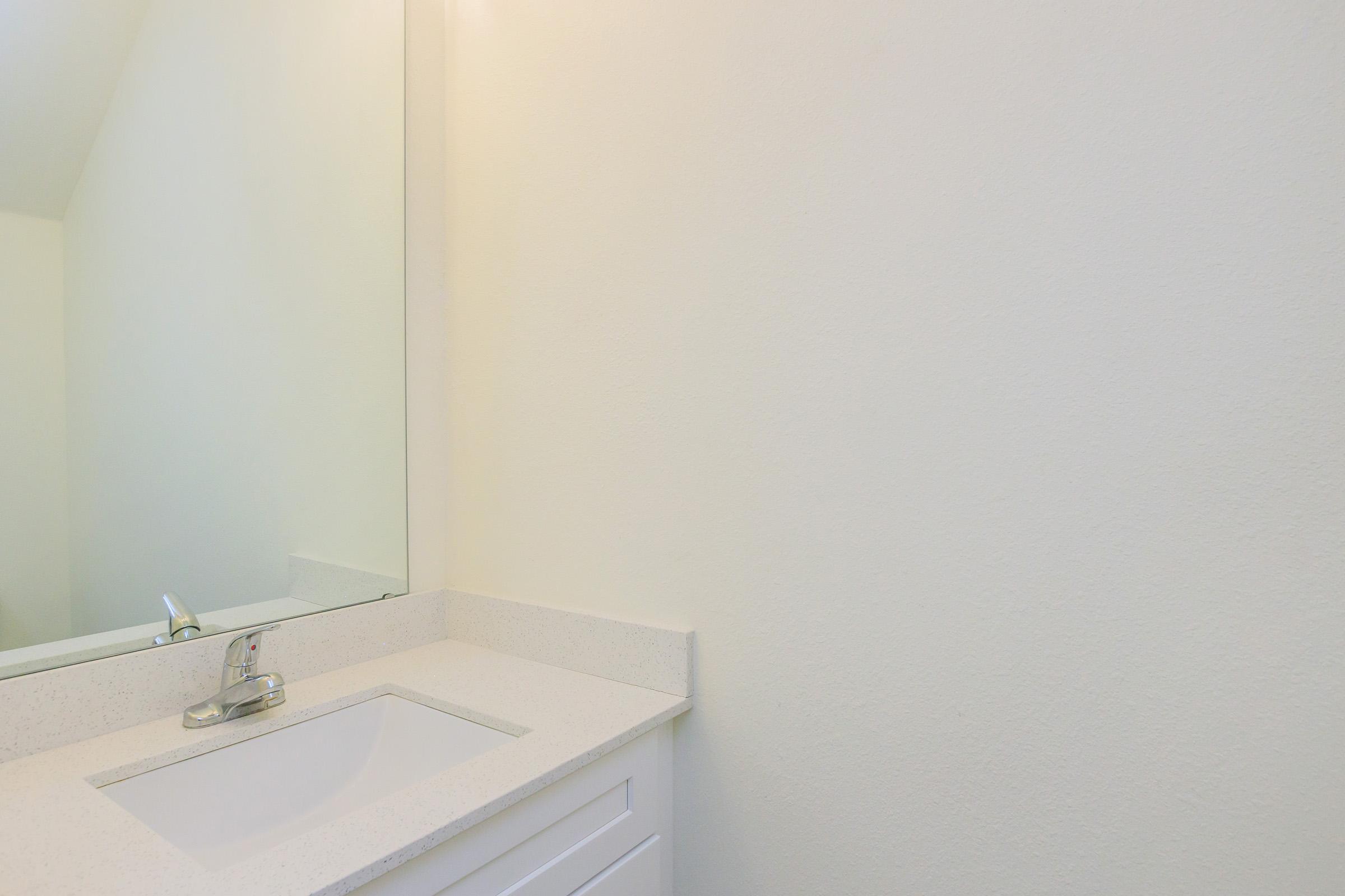 A clean and minimalistic bathroom vanity area featuring a white sink, a simple faucet, and a large mirror above the sink. The walls are painted a soft white, creating a bright and airy atmosphere. There are no visible decorations or personal items, emphasizing a tidy aesthetic.