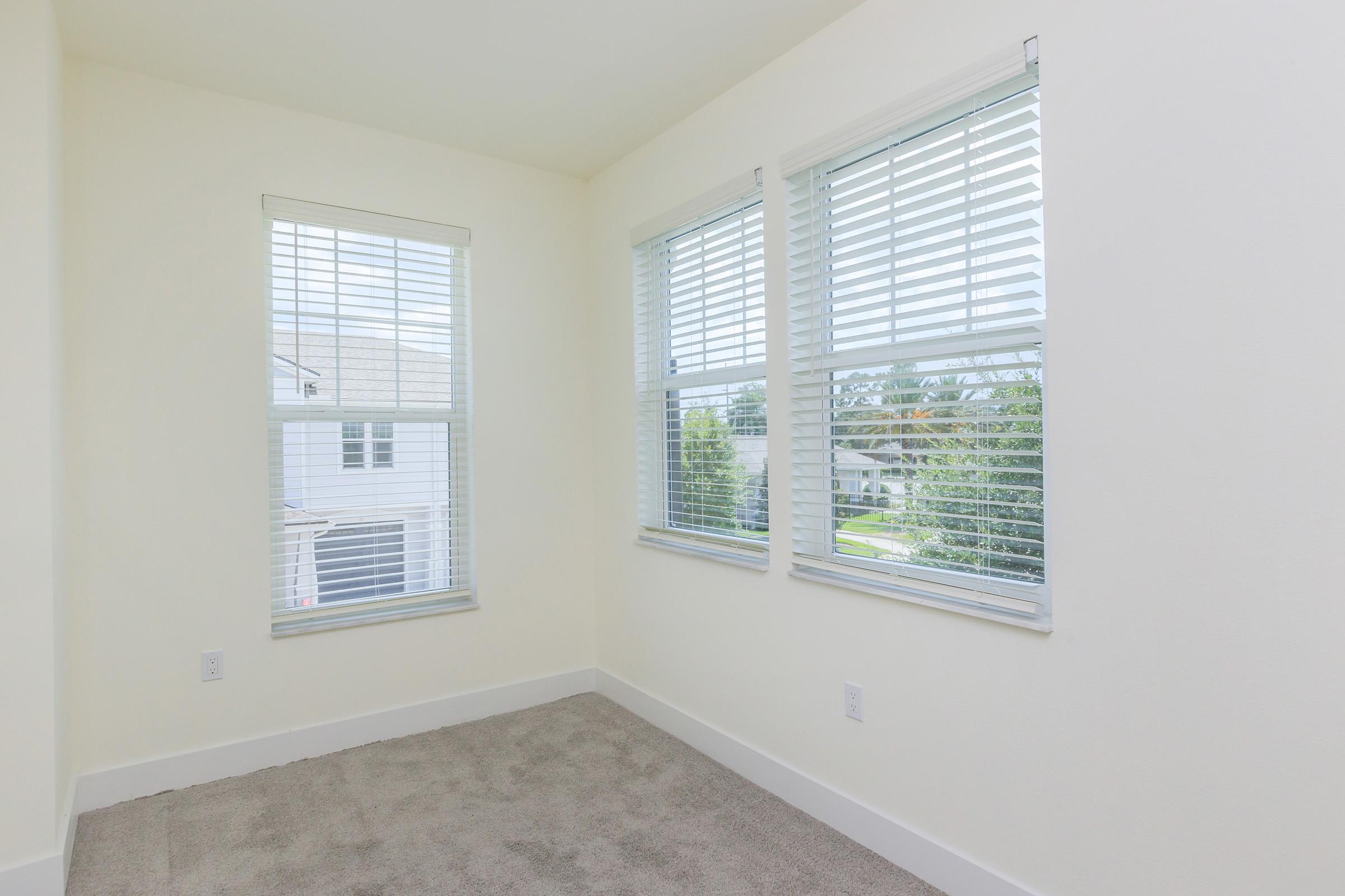 Bright, empty room featuring three windows with white blinds. The walls are painted light yellow, and the floor is covered with a soft, light gray carpet. Outside the windows, residential buildings and greenery are visible. The overall space is clean and inviting, suitable for various uses.