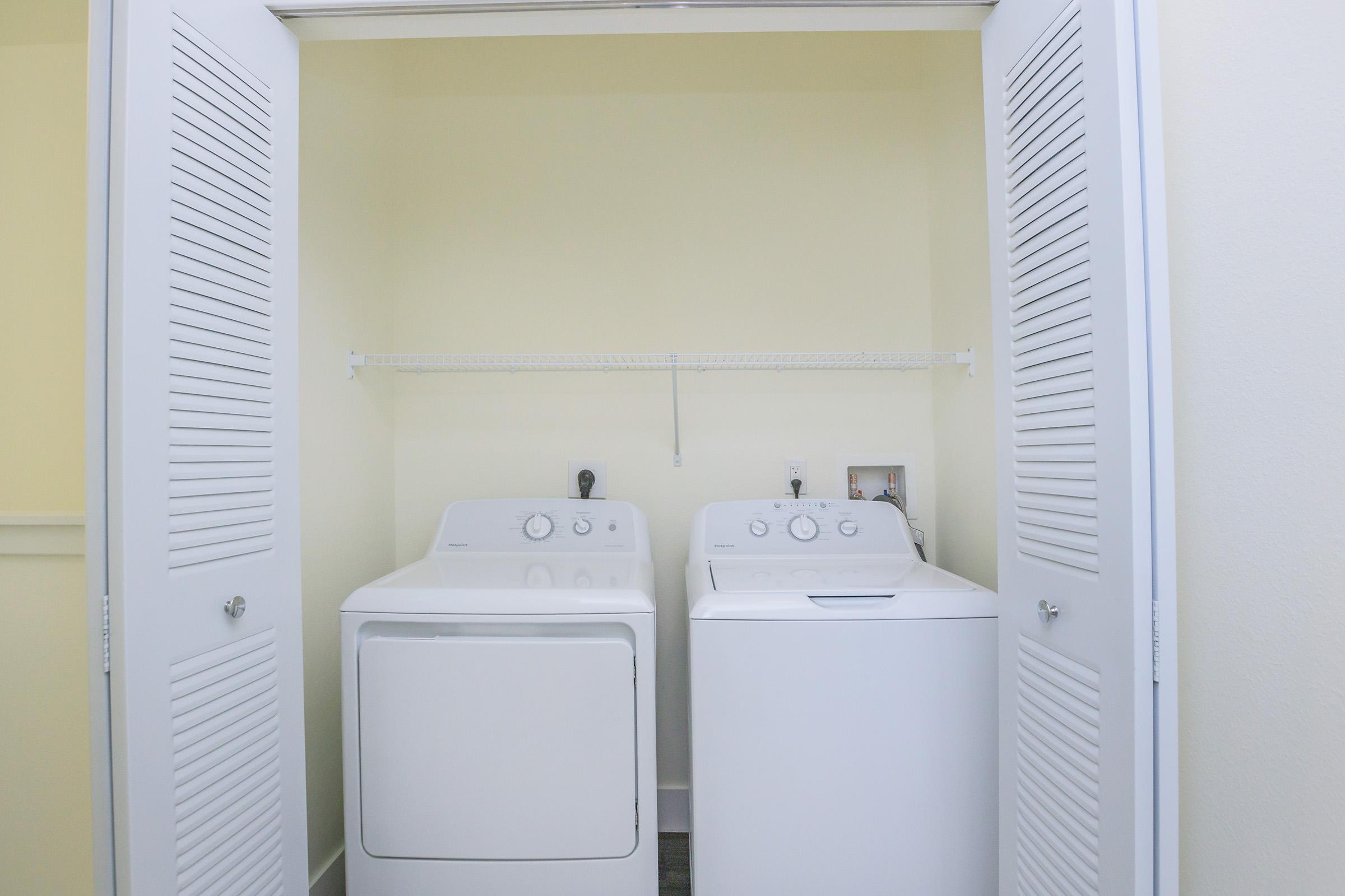 A laundry room featuring a stacked washer and dryer set, arranged side by side, with a shelf above for storage. The walls are painted a light yellow, and the space is neatly organized with open double doors revealing the appliances.
