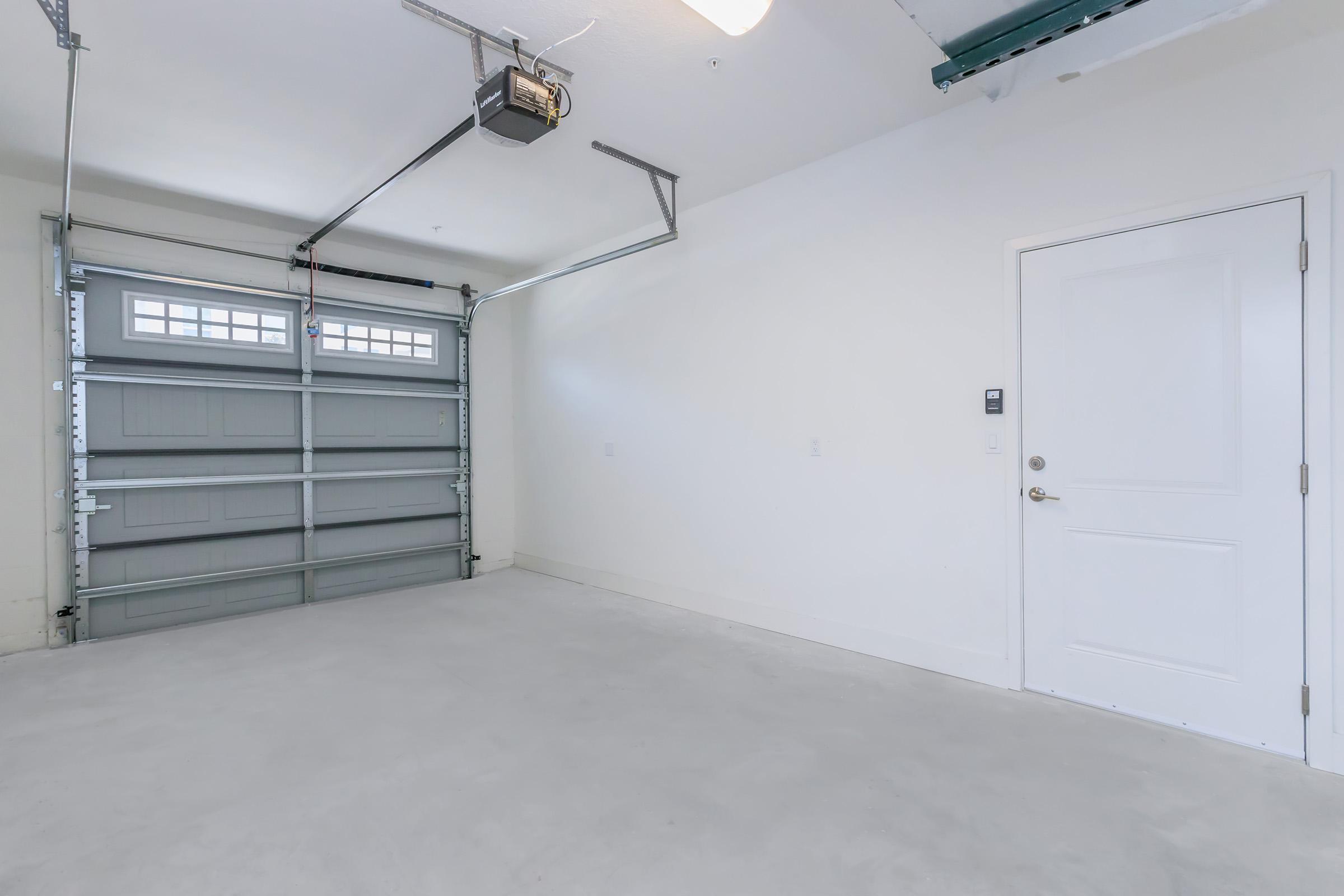 A clean and empty garage with a gray overhead door partially open, white walls, a concrete floor, and a closed door on the right side. The space is well-lit, showcasing a simple and organized layout, perfect for storage or use as a workspace.