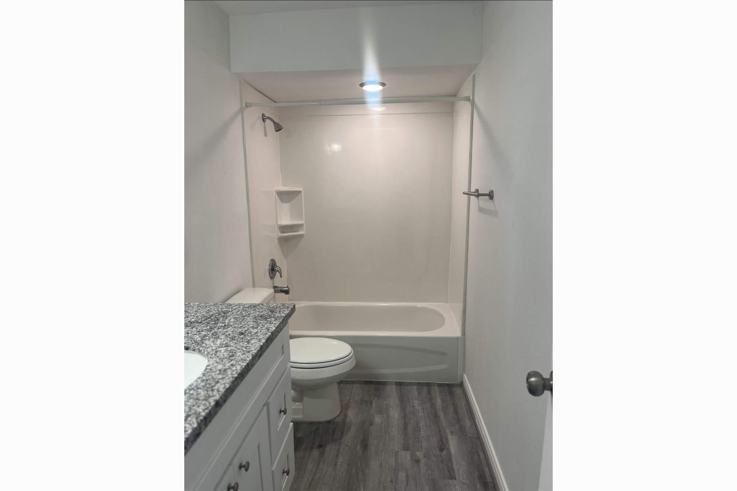 A clean and modern bathroom featuring a white bathtub/shower combination, a small shelving unit in the corner, a granite countertop with a sink, and a toilet. The walls are painted white, and the floor is a light-colored laminate, providing a bright and spacious appearance.