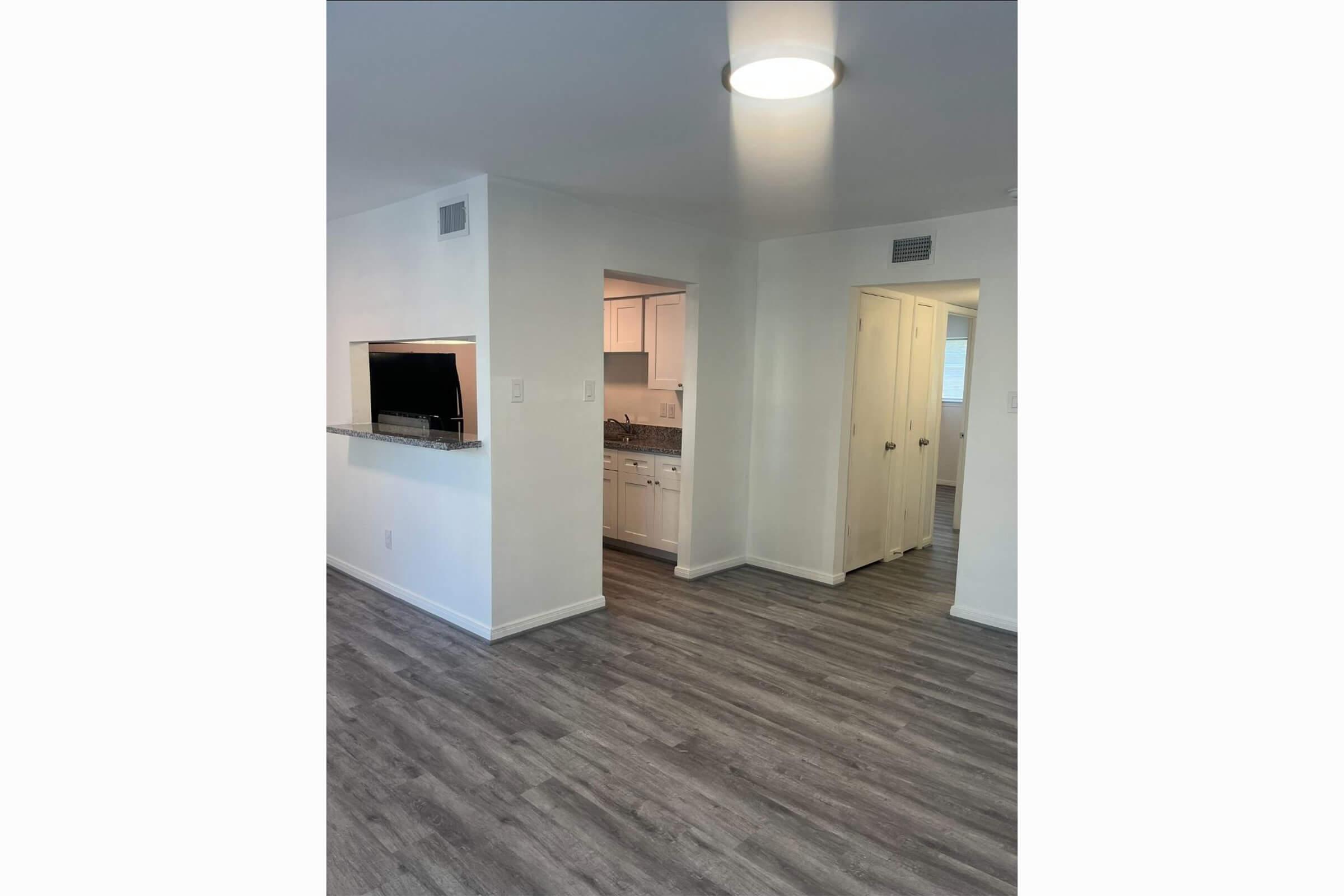 A bright, modern living space featuring light-colored walls and gray laminate flooring. The room includes a small opening leading to a kitchen area with white cabinetry. Doors lead to additional rooms. A ceiling light provides illumination.