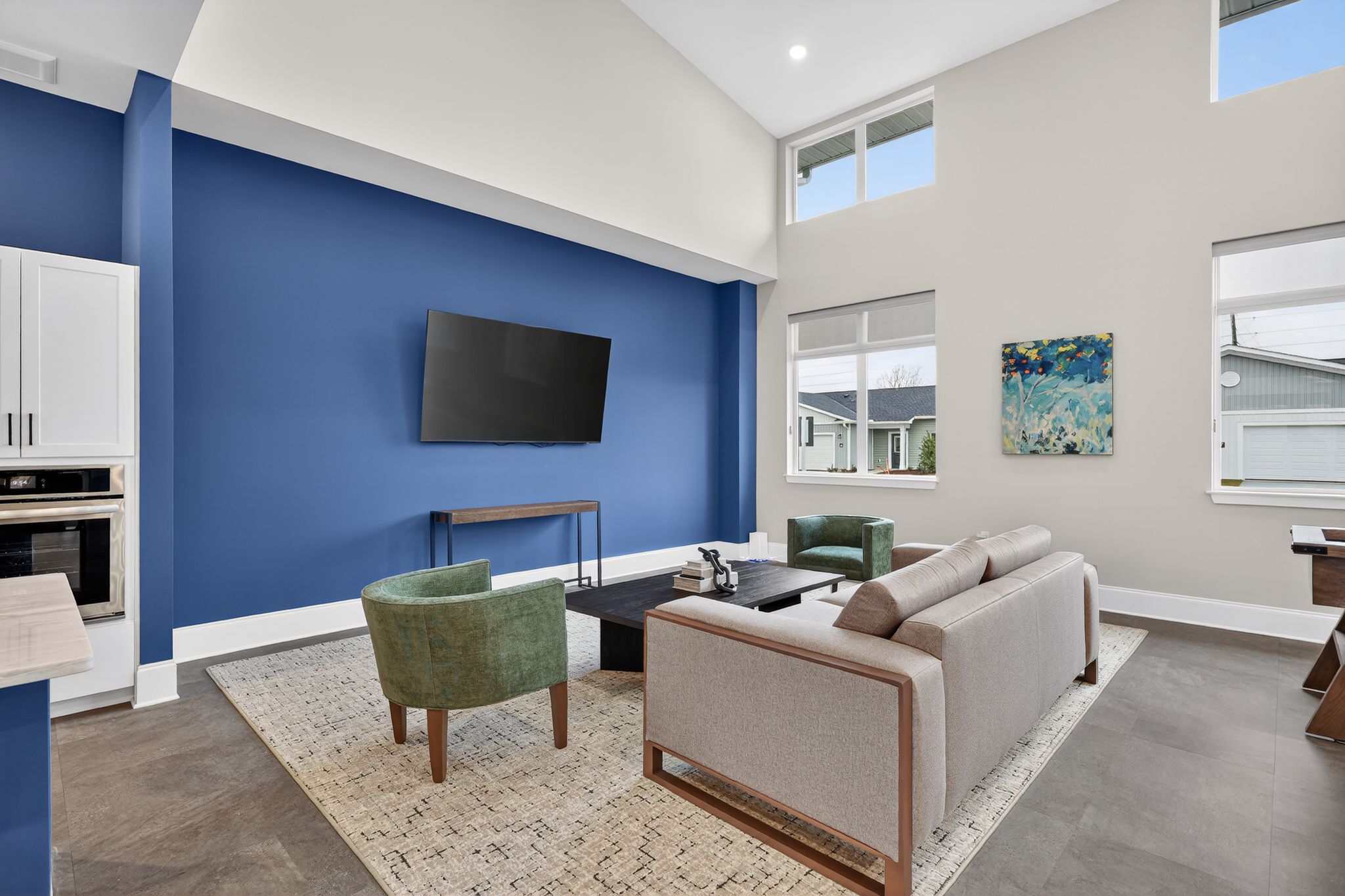 A modern living room featuring a high ceiling and a bold blue accent wall. There's a large flat-screen TV mounted on the wall, a light-colored sofa, and two green chairs around a dark coffee table. Natural light filters through large windows, illuminating a contemporary rug underfoot.