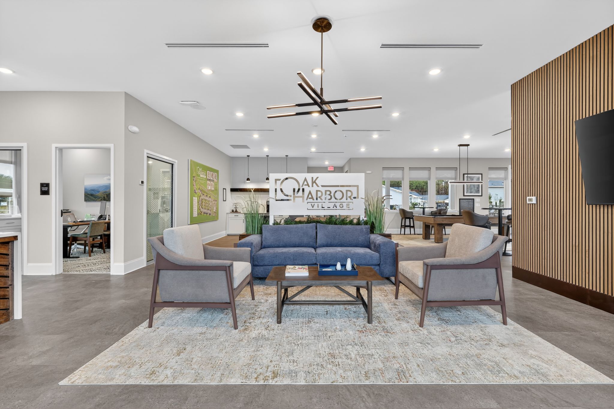 A modern interior lounge area featuring a blue sofa, two armchairs, and a coffee table on a light rug. There are decorative plants, contemporary light fixtures, and artwork on the walls, creating a welcoming atmosphere in a community space.