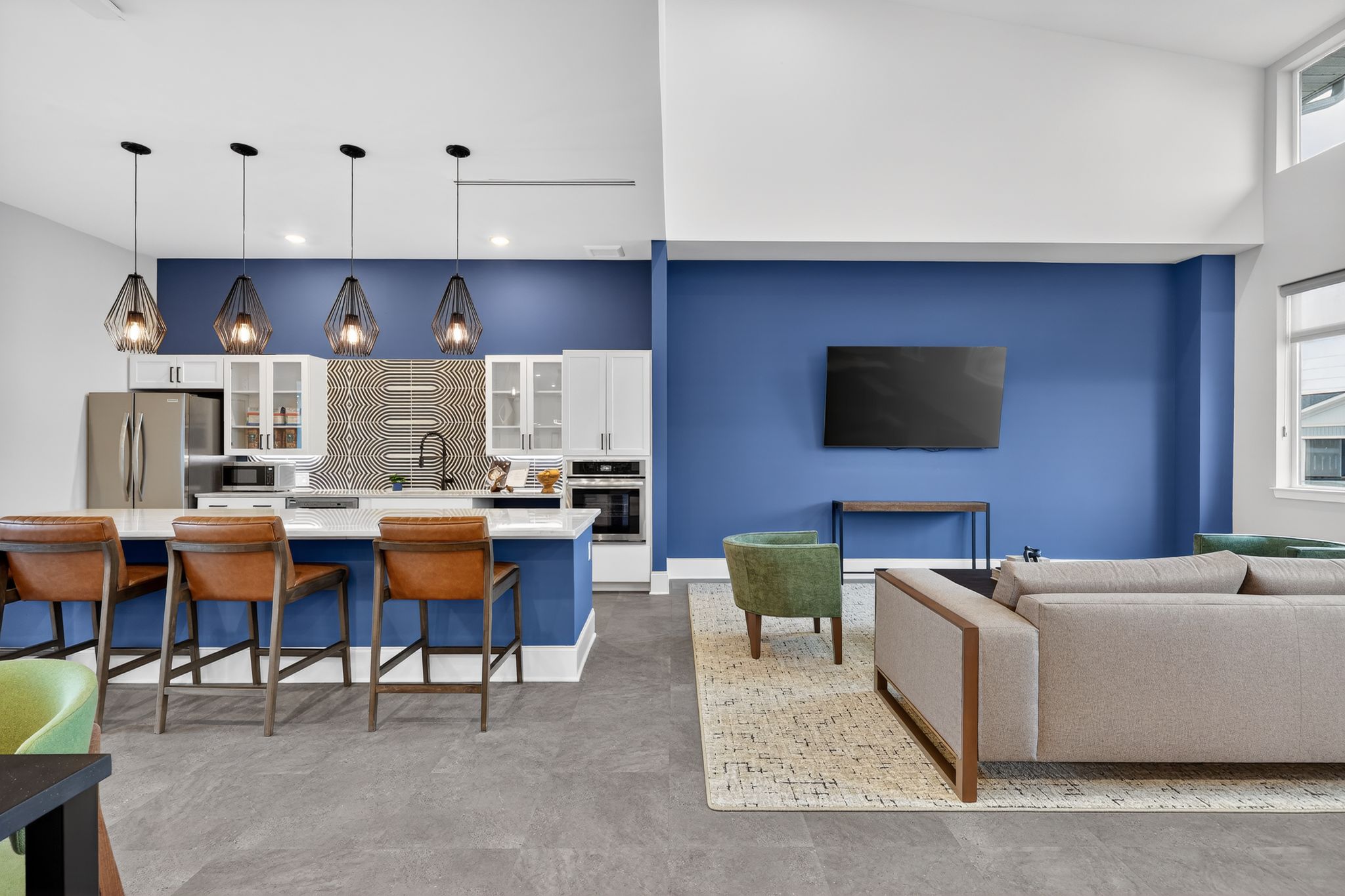 Modern kitchen and living area featuring a large island with bar stools, white cabinetry, and stainless steel appliances. The blue accent wall contrasts with neutral tones. A flat-screen TV is mounted above a wooden table, while a cozy sofa and green chairs add comfort to the space. Large windows allow natural light.