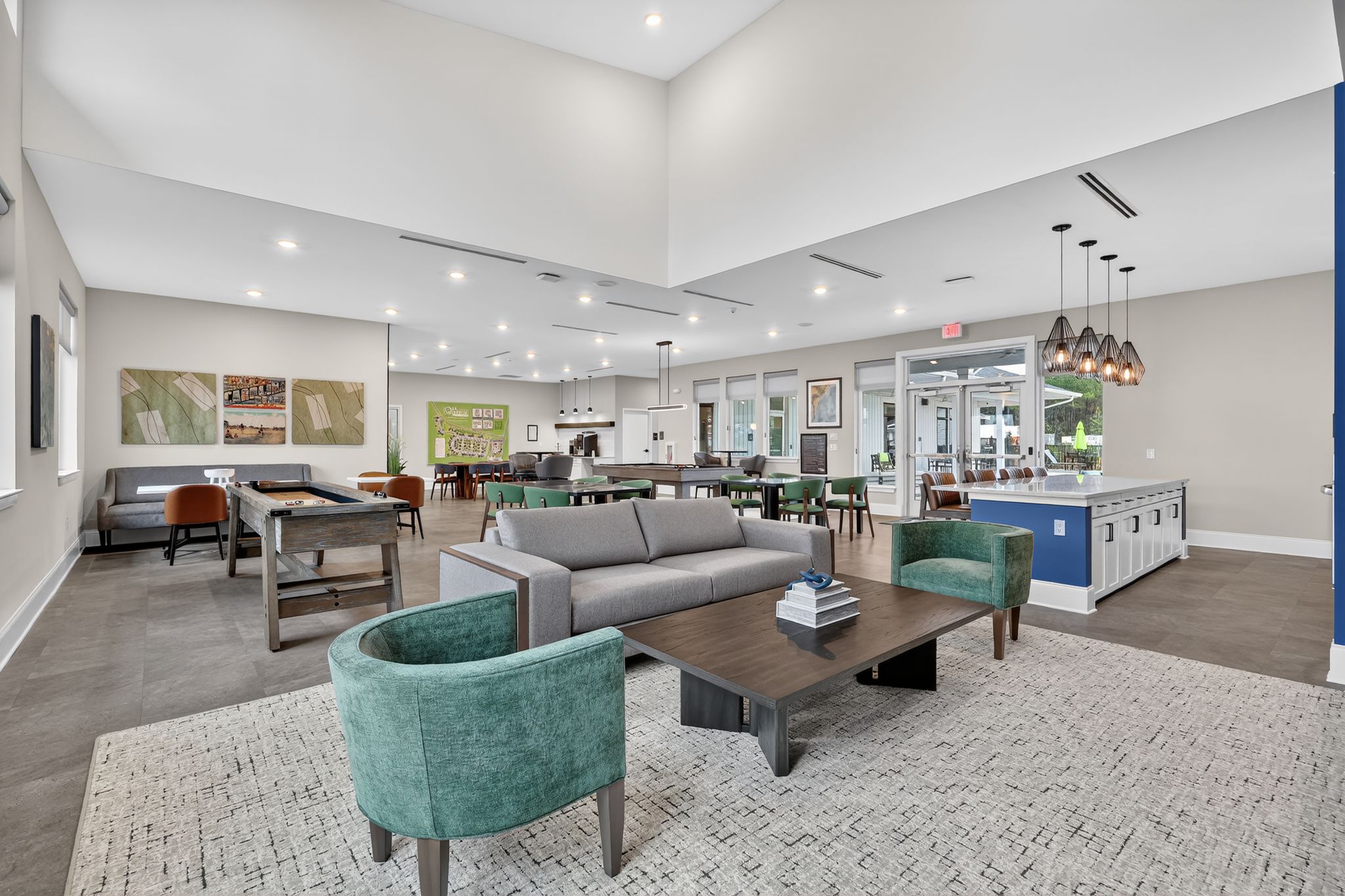 A modern communal lounge area featuring a gray sofa and green accent chairs around a dark coffee table. There's a light fixture with multiple bulbs overhead, and large windows provide natural light. The space includes dining tables and a kitchenette area visible in the background.