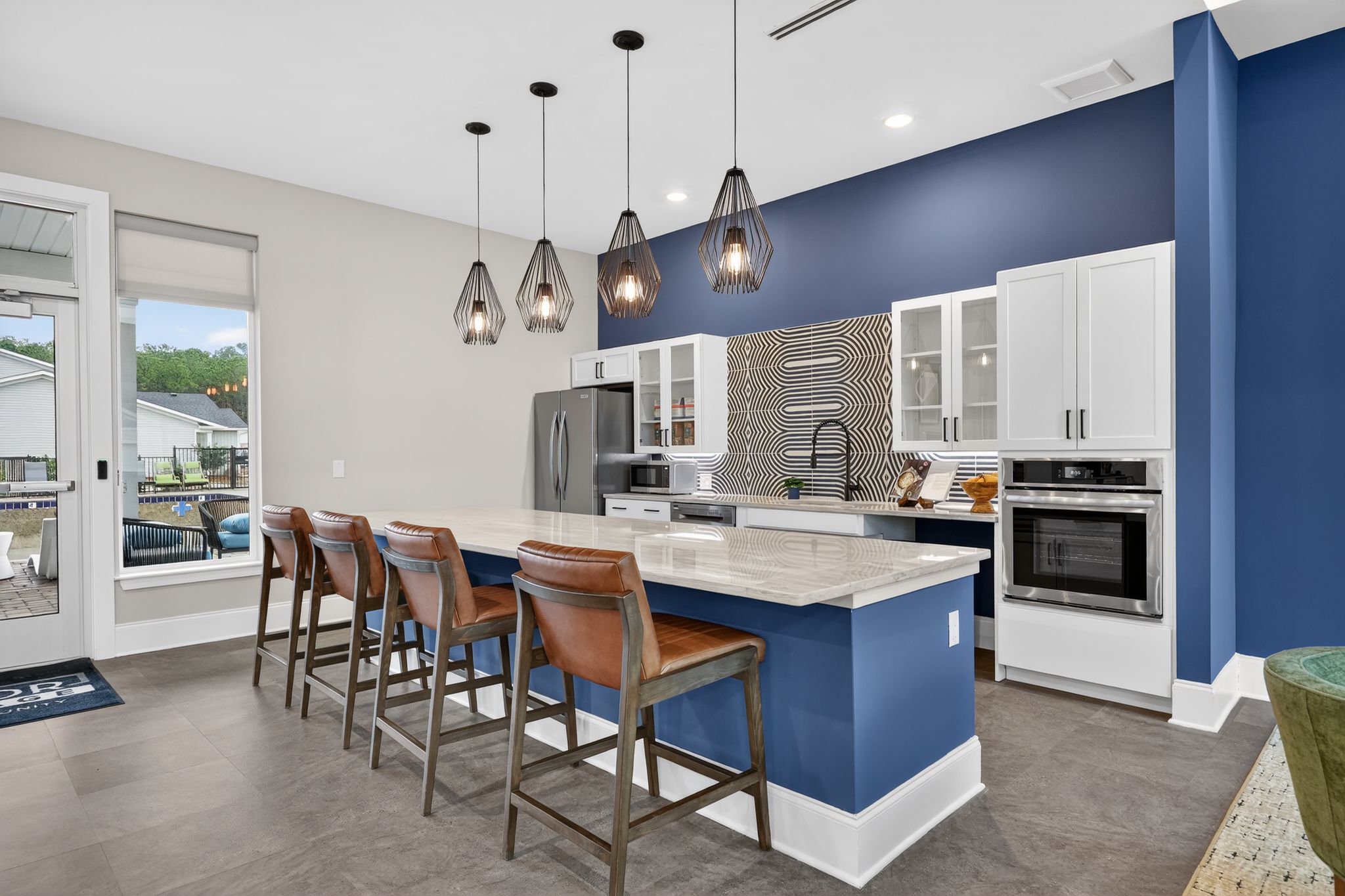 Modern kitchen featuring a large island with bar seating, pendant lights, and navy blue accent wall. Equipped with stainless steel appliances, white cabinetry, and a stylish backsplash. Large windows provide natural light and a view of the outdoor space. 