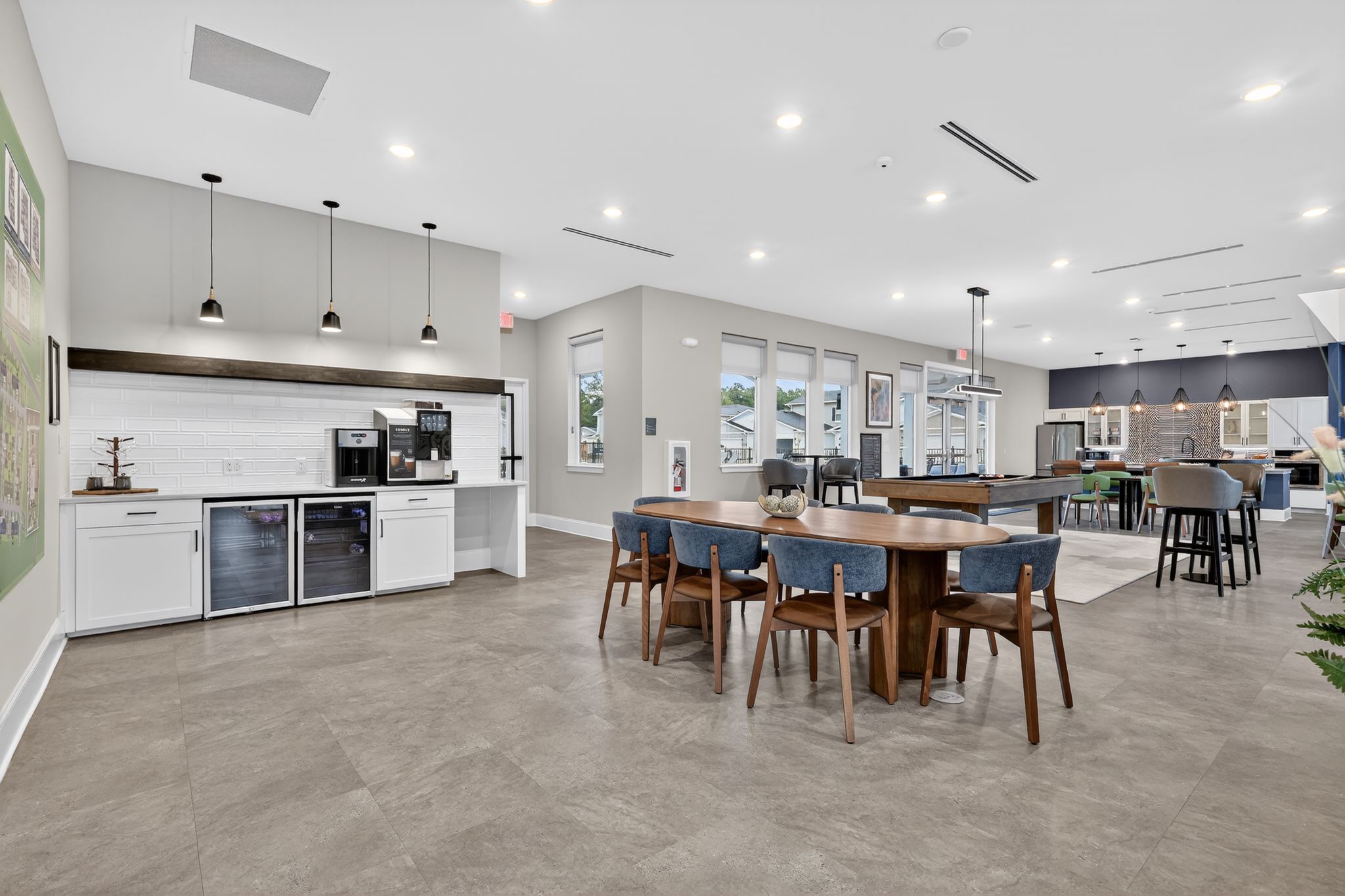 Modern communal space featuring a large dining table with blue upholstered chairs, a kitchenette with a coffee machine and beverage cooler, and ample natural light from large windows. The decor includes neutral tones and contemporary lighting, creating an inviting atmosphere for social gatherings.