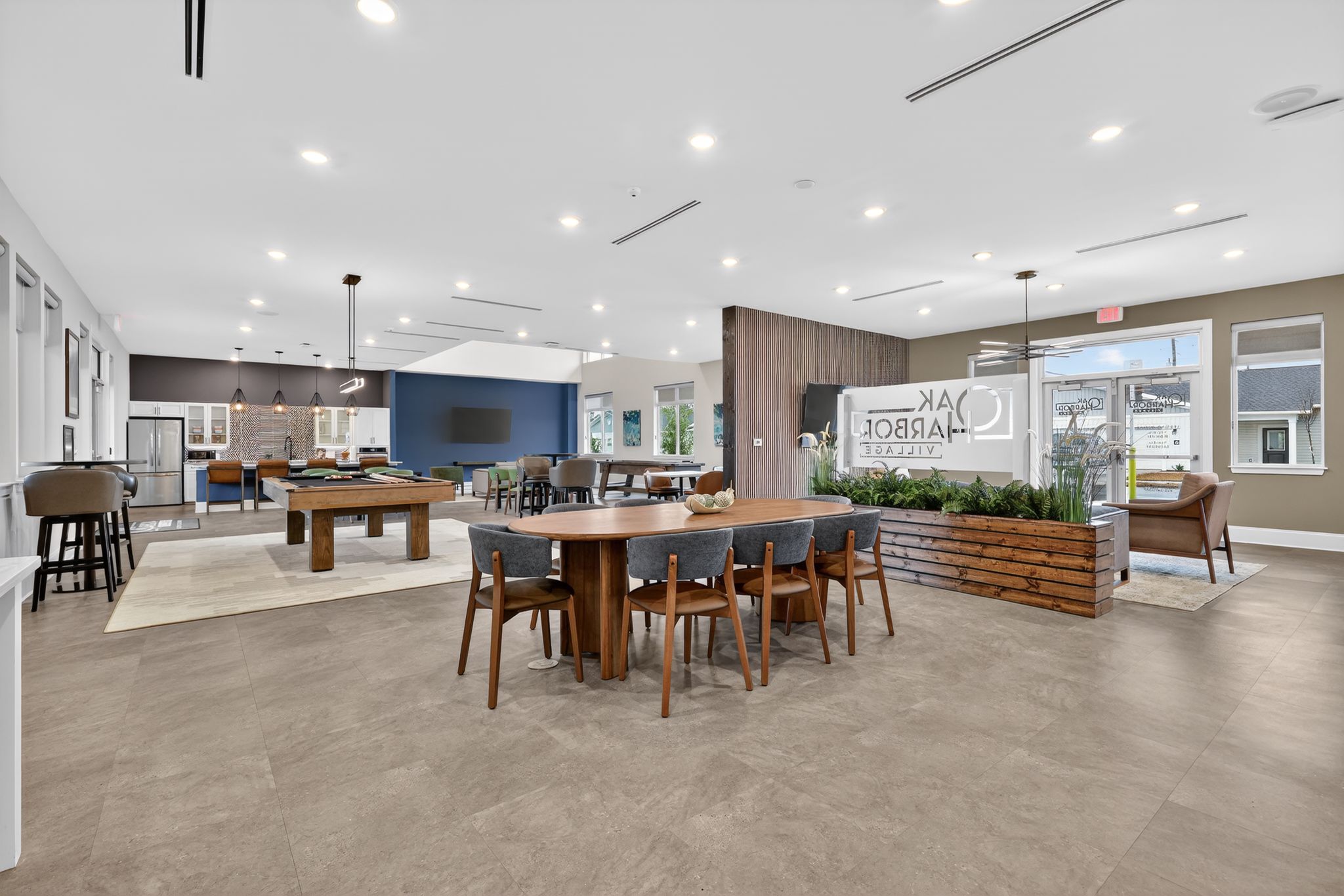 Modern indoor space featuring a welcoming lounge area with seating, a dining table, and a pool table. Large windows let in natural light, and there are decorative plants and a sign for "Oak Arbor Villas." The design is contemporary with an open layout, ideal for socializing and relaxation.