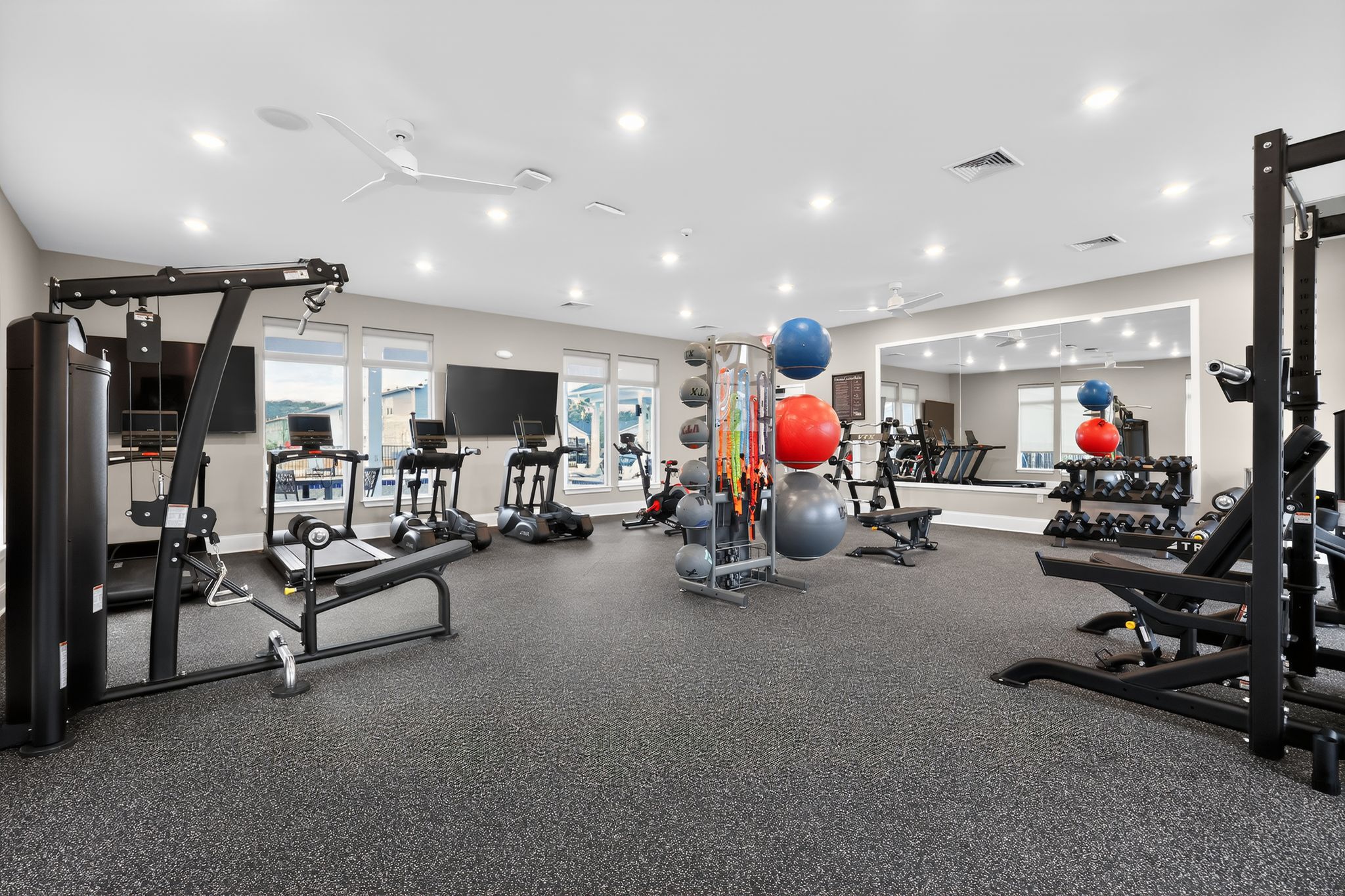 A modern gym space featuring various exercise equipment, including treadmills, weight machines, and free weights. The room is well-lit with large windows, and colorful exercise balls are arranged in an organized manner, creating an inviting atmosphere for workouts.
