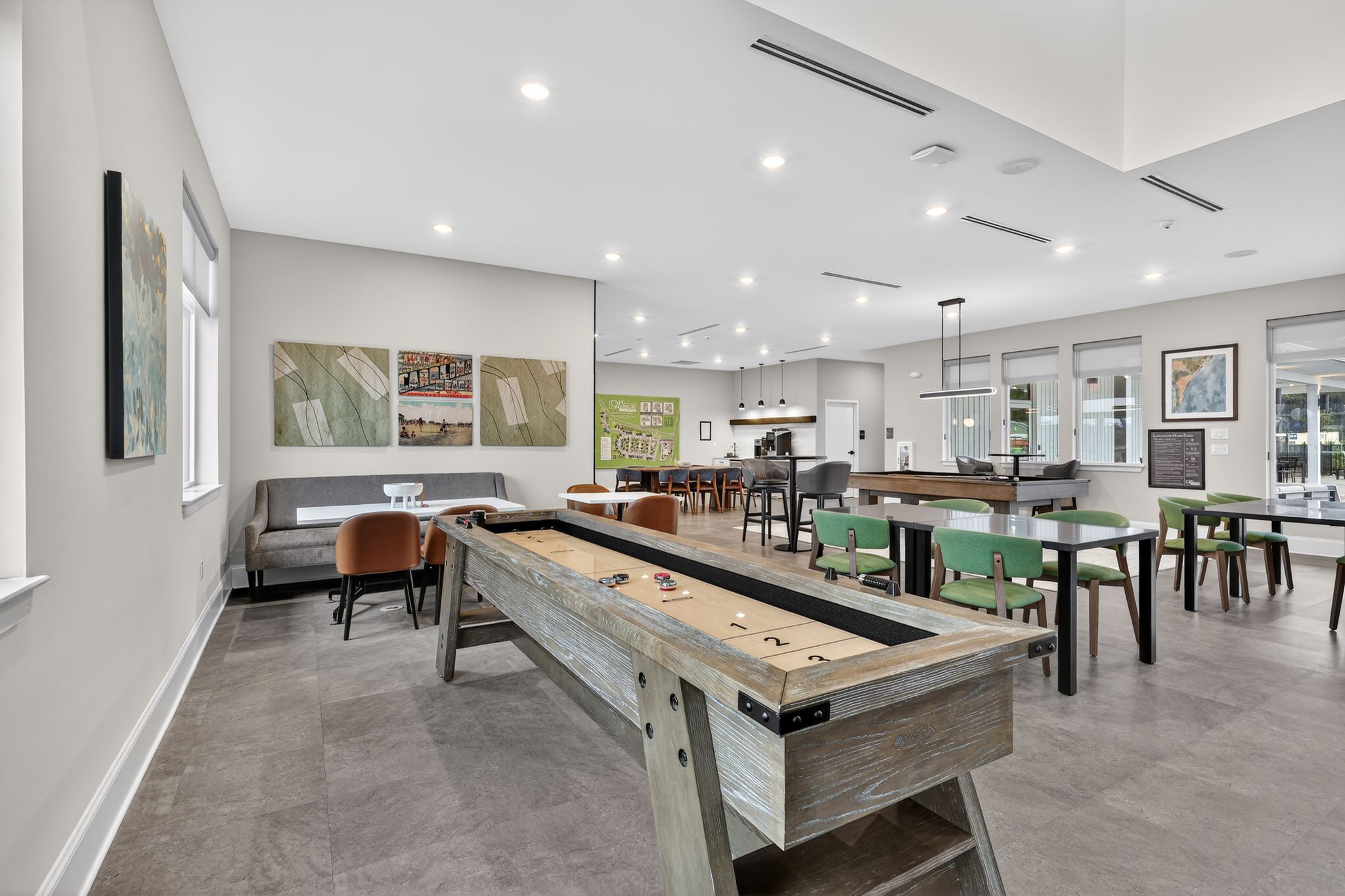 A modern lounge area featuring a shuffleboard table, comfortable seating, and large windows. The walls are decorated with artwork, and there are tables with chairs. The space has a bright, welcoming atmosphere, ideal for socializing or relaxation.