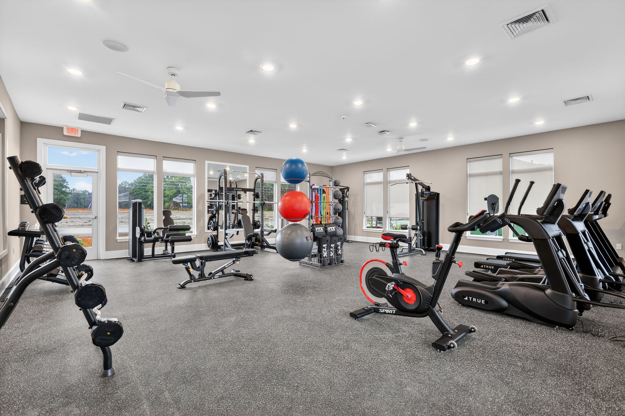 A modern gym interior featuring various exercise equipment, including dumbbells, weight machines, elliptical trainers, and exercise balls. Large windows allow natural light, and the flooring is dark, providing a clean and bright workout environment.