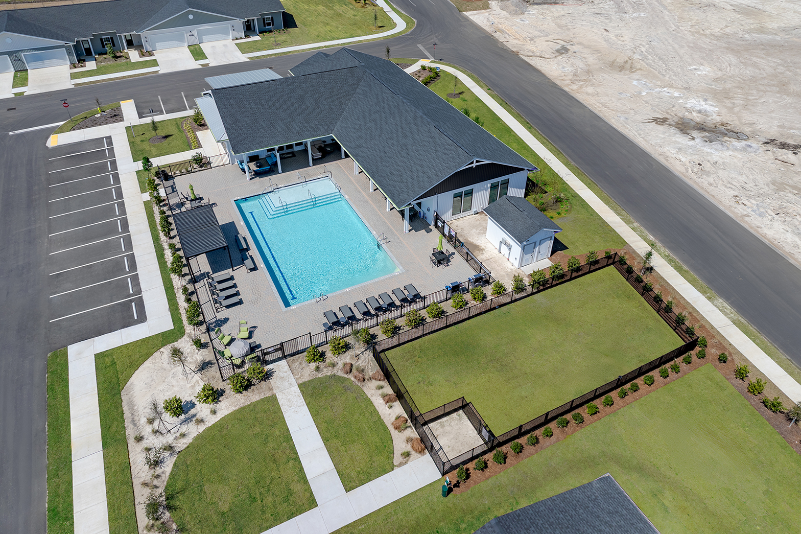 An aerial view of a modern community with a swimming pool, lounge chairs, and a clubhouse. Surrounding the pool are landscaped areas and parking spaces. The image shows green lawns, pathways, and under-construction lots in the background, indicating the development of a residential area.