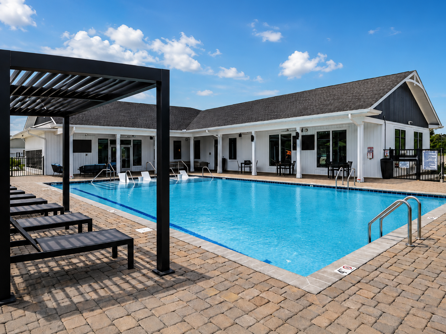 A spacious outdoor swimming pool surrounded by lounge chairs, with a pergola on the left. In the background, there is a modern white building with large windows and a dark roof, set against a clear blue sky with a few clouds.