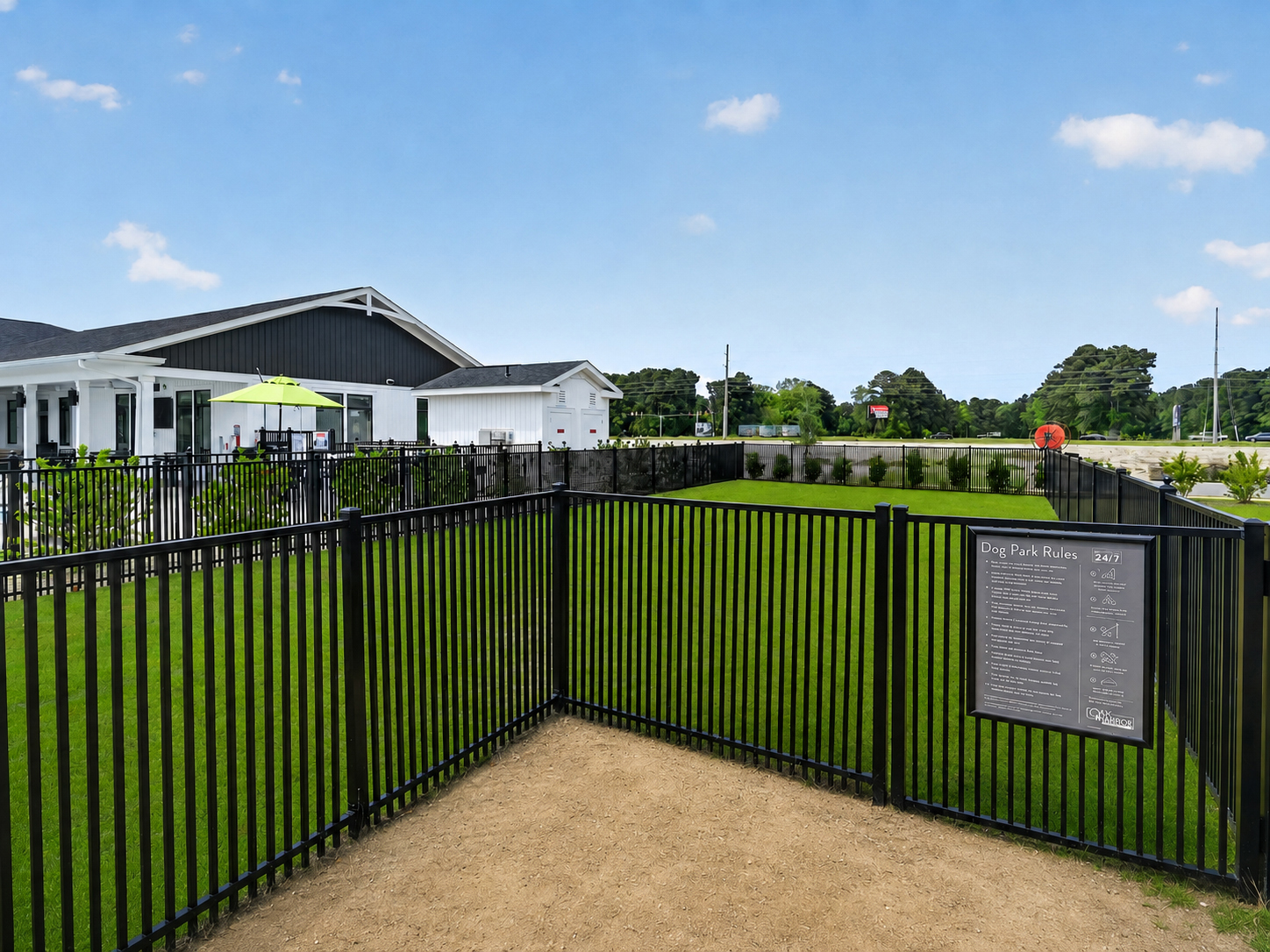 A dog park enclosed by black metal fencing, featuring a sandy area and well-maintained green grass. In the background, a modern building with a covered sitting area is visible. A sign detailing dog park rules is mounted on the fence. Clear blue sky adds to the outdoor atmosphere.