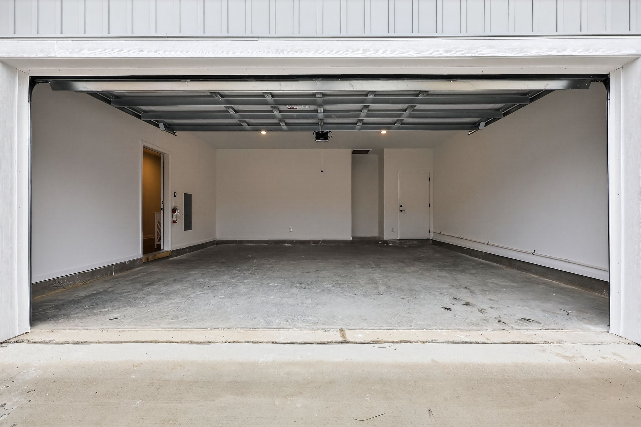 Empty garage space with a concrete floor, white walls, and overhead lighting. The garage door is open, revealing the interior, which includes a side door and a wall-mounted electrical panel. The area is clean and unobstructed, suitable for parking or storage.