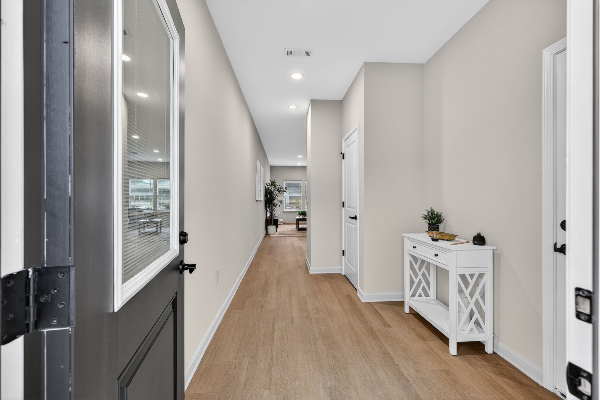 A modern hallway featuring light-colored walls and wooden flooring. On the left, a black door opens to the outside. A white side table with a small plant sits against the wall. The hallway leads to a well-lit space in the background with plants, suggesting a welcoming atmosphere.