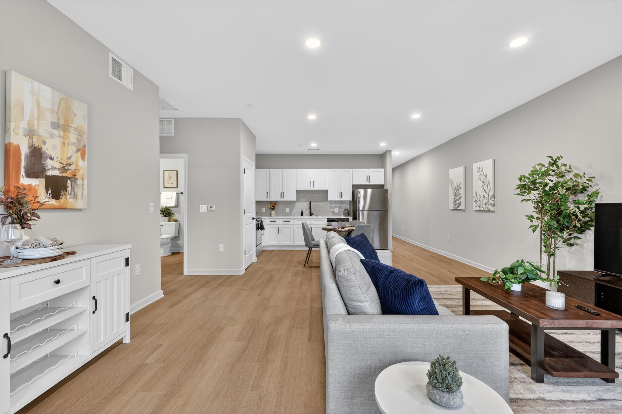 Modern living room and kitchen area featuring light-colored walls, hardwood flooring, and contemporary furniture. The living space includes a grey couch, a coffee table, and decorative plants. The kitchen is equipped with white cabinets and stainless steel appliances, creating a bright and inviting atmosphere.
