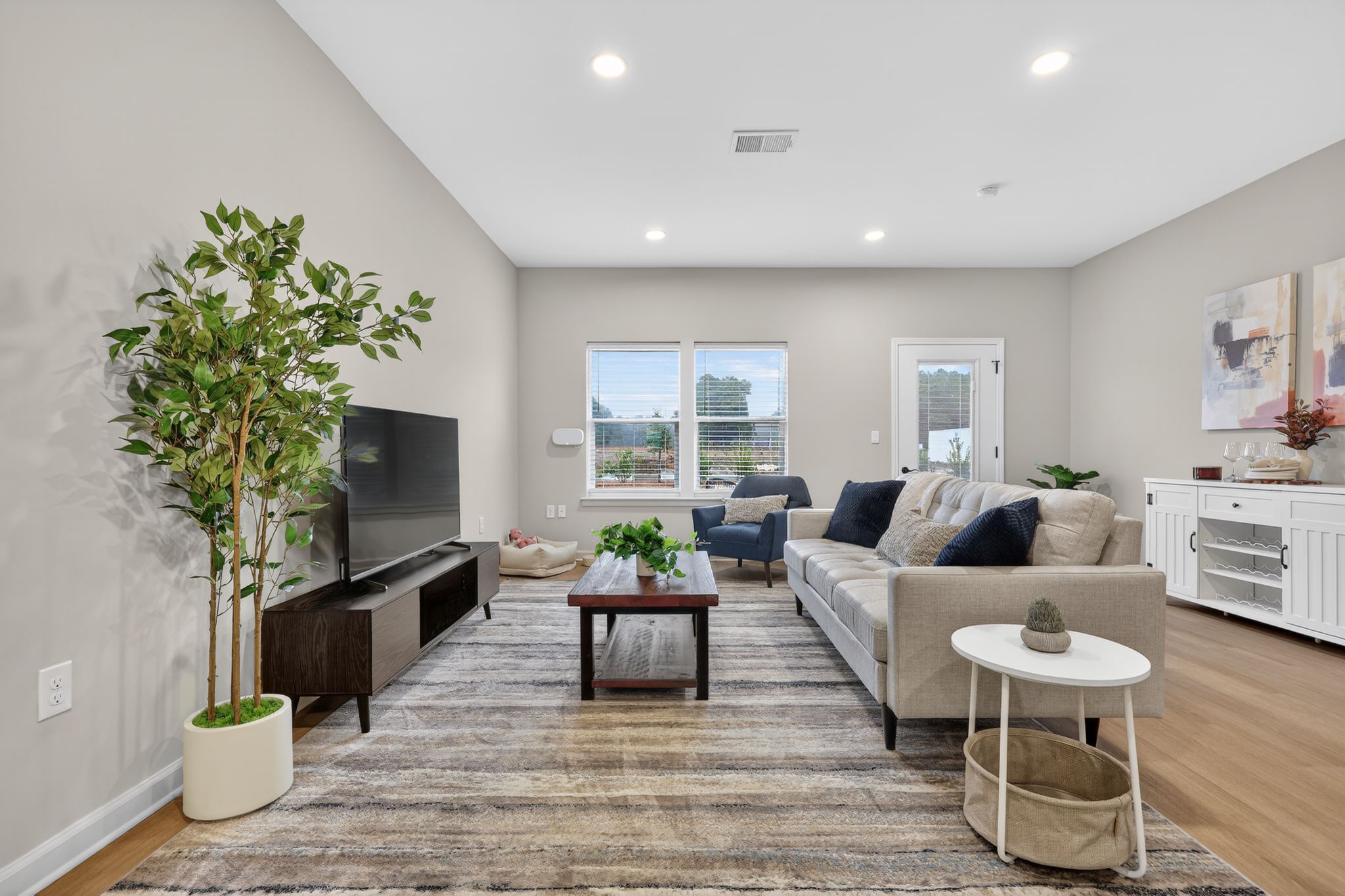 Modern living room featuring a comfortable beige sofa, a dark wood TV stand with a television, and a light-colored area rug. A potted plant adds a touch of greenery, while natural light filters through two windows. Decorative items are arranged on a white sideboard against the wall.