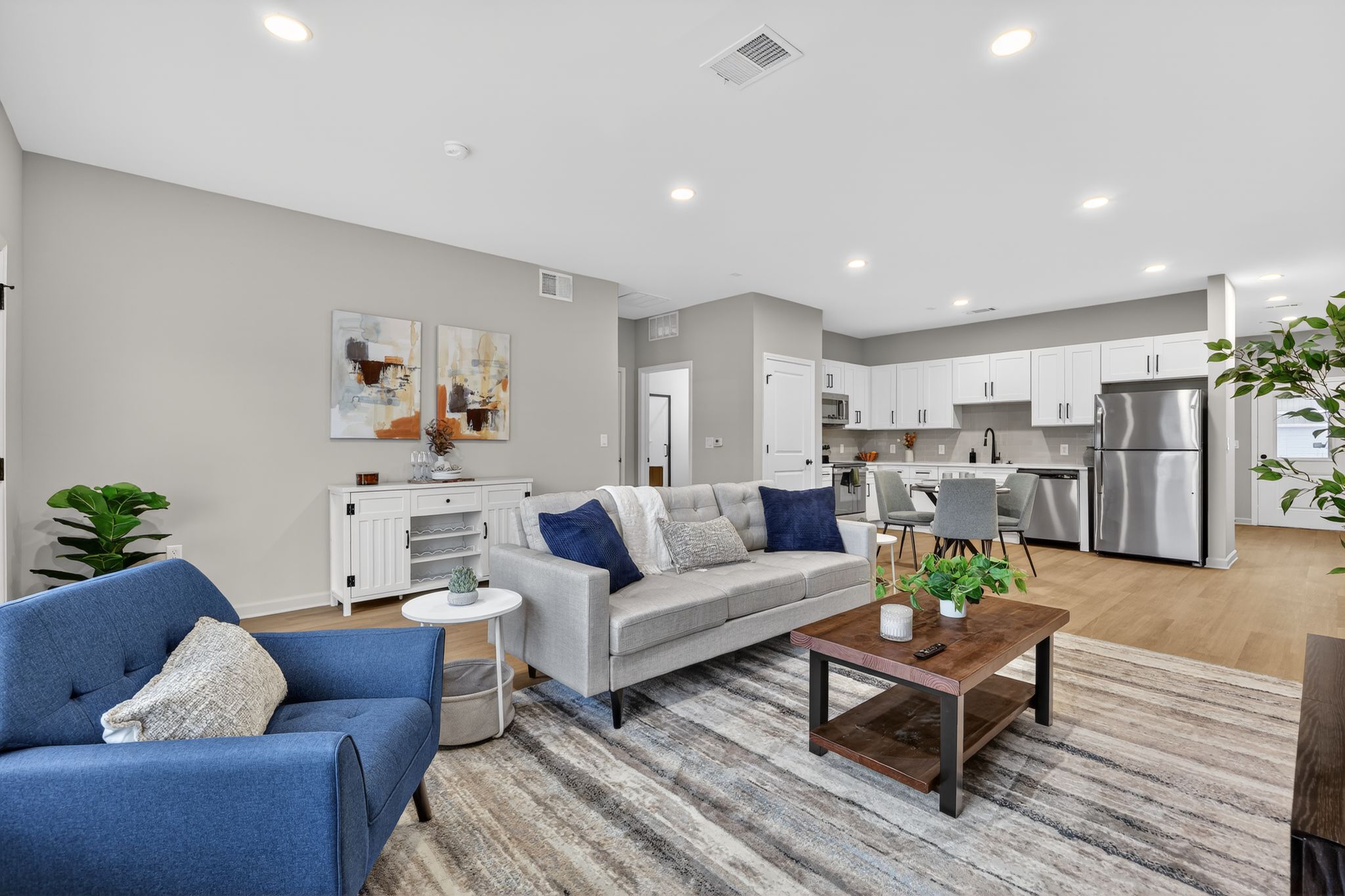 A modern living room featuring a blue and gray sofa, a wooden coffee table with decorative items, and a plush area rug. The space includes a dining area with a kitchen visible in the background, white cabinetry, and stainless steel appliances. Light-colored walls and ample lighting create a bright, inviting atmosphere.