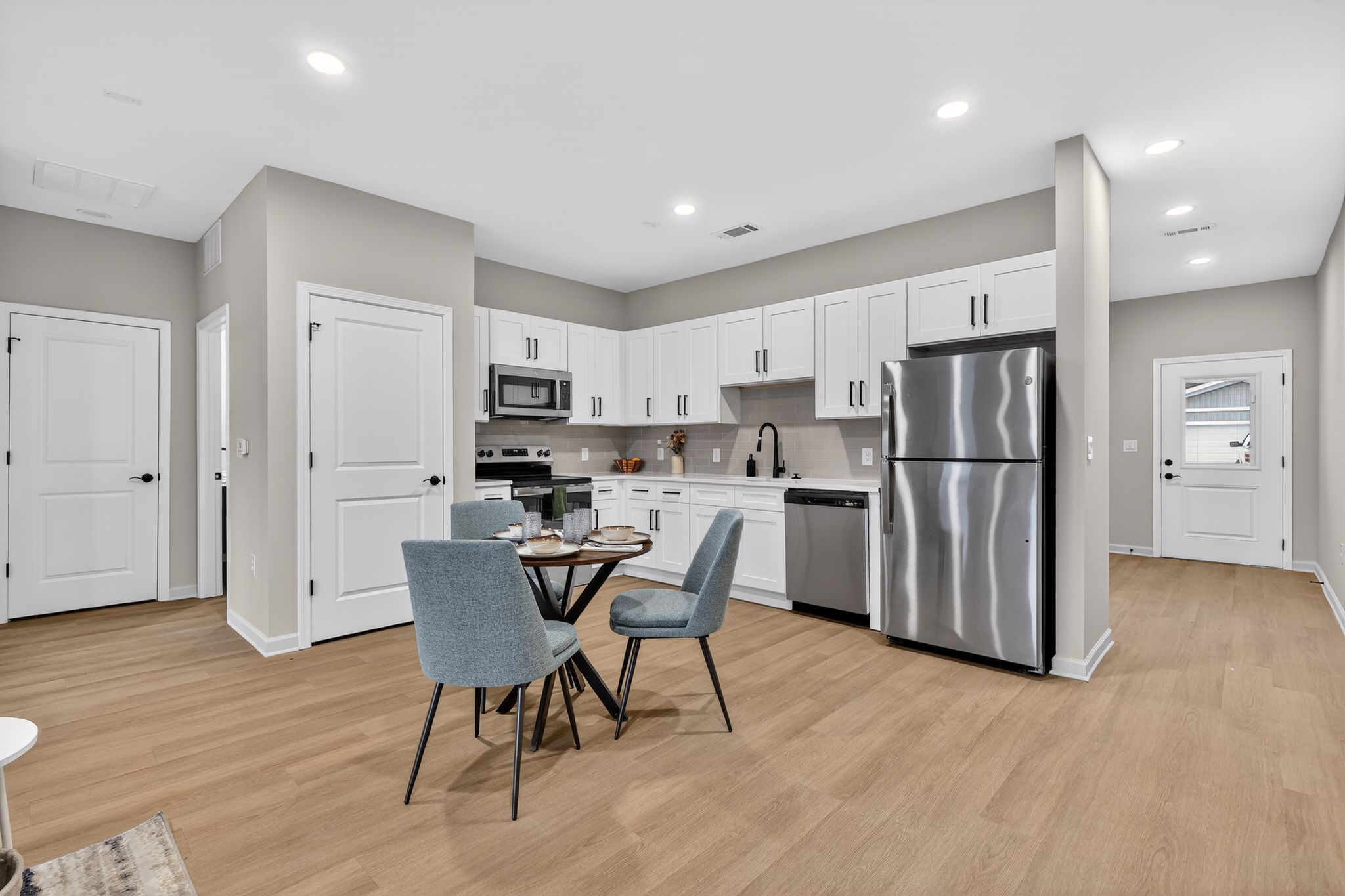 Modern kitchen with light gray walls and white cabinetry. Features stainless steel appliances including a microwave, refrigerator, and dishwasher. A round dining table with two blue chairs sits in the center, and there are two doors leading to other rooms. Bright, well-lit space with wooden flooring.