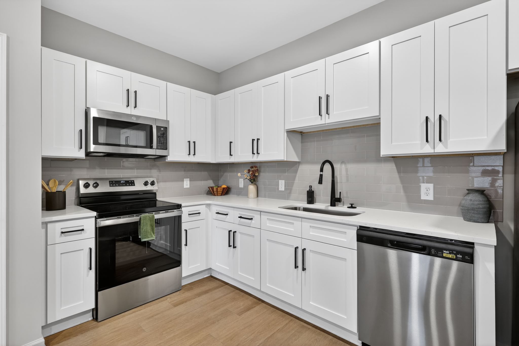 Modern kitchen featuring white cabinets, stainless steel appliances, a black stove with an oven, a microwave above, and a dishwasher. The countertop is light-colored with a gray tile backsplash. A sink with a black faucet is visible, along with decorative items in a bowl and a vase. The flooring is light wood.