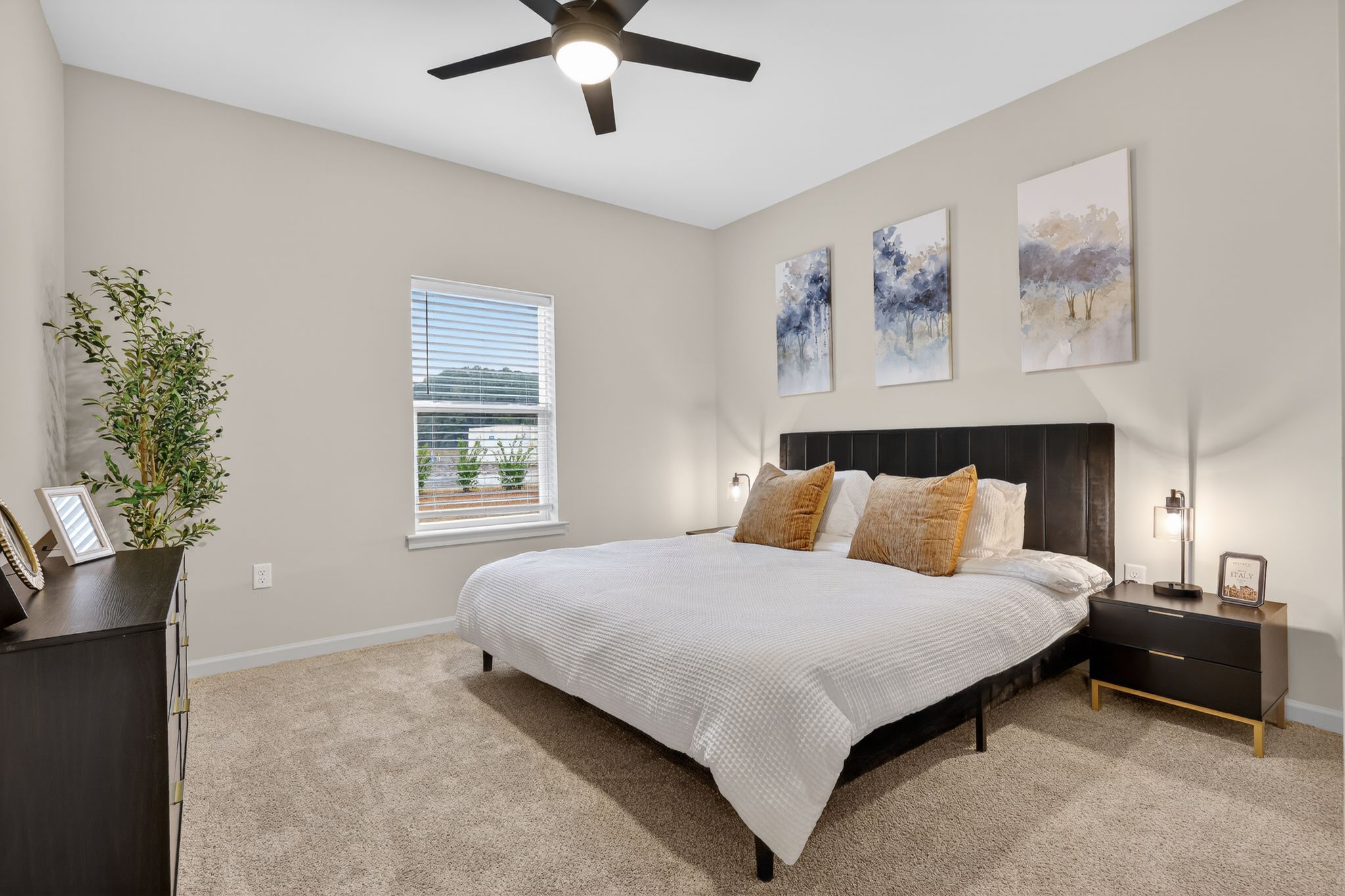 A cozy bedroom featuring a large bed with white bedding and two decorative pillows. The walls are painted in a soft color, and there are three framed artworks above the bed. A bedside table holds a lamp, and a dresser is visible to the left. Natural light floods the room through a window with blinds.