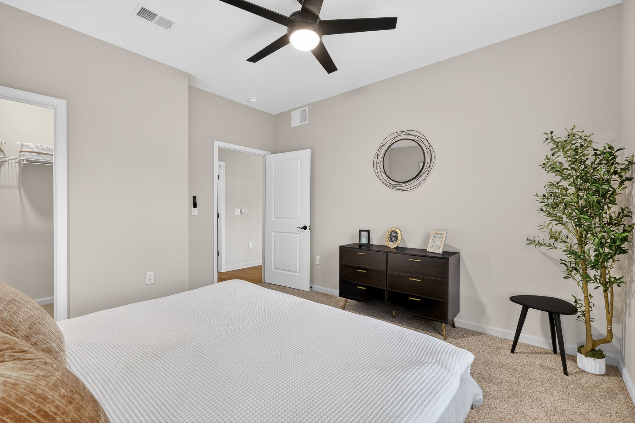 A well-lit bedroom featuring a queen-sized bed with a white quilt, a dark wood dresser against the wall, a small black accent table, and a decorative plant in the corner. The walls are painted a soft beige, and there is a modern ceiling fan. A closet and doorway are visible in the background.