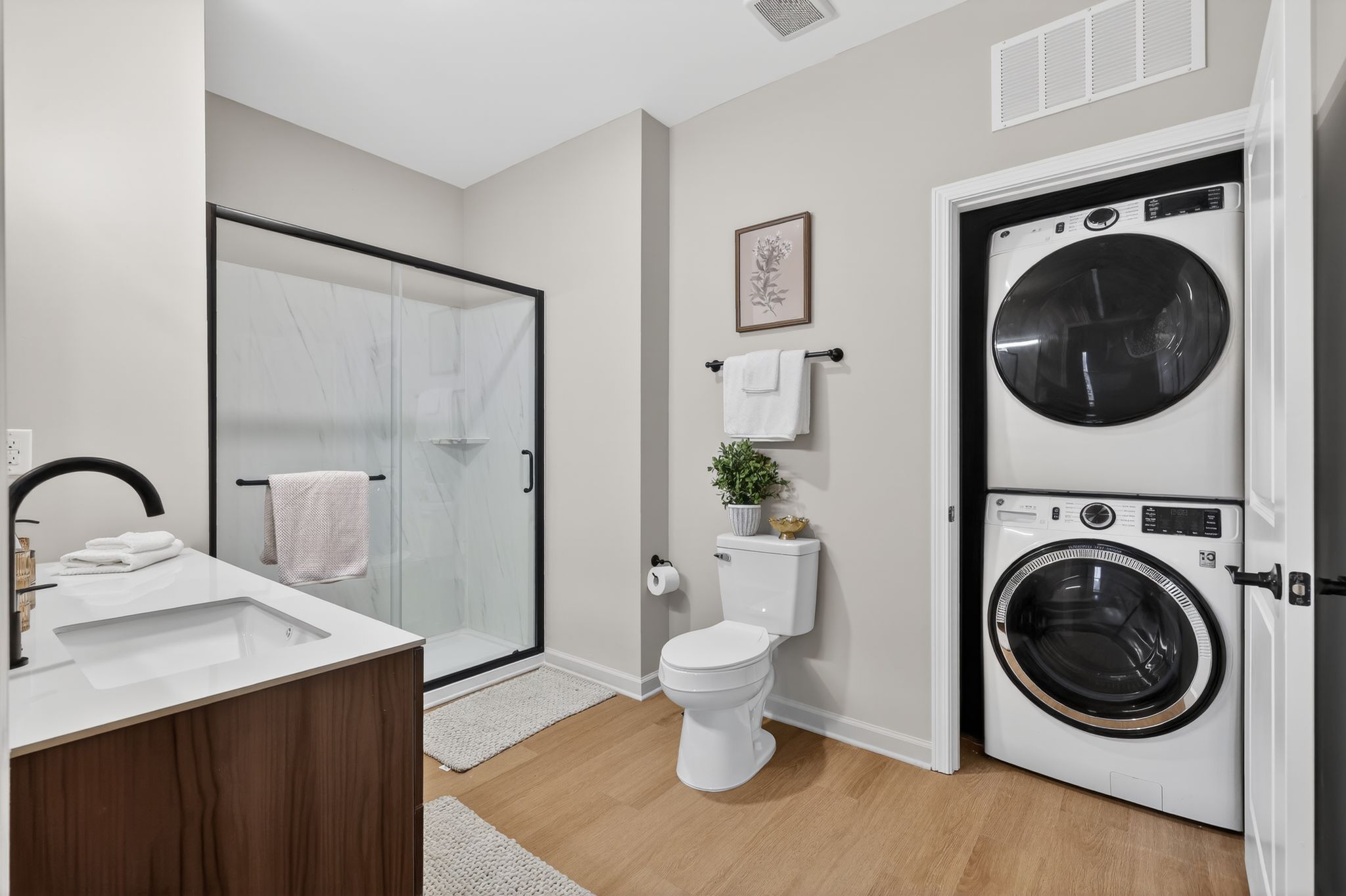 A modern bathroom featuring a walk-in shower with glass doors, a sleek vanity with a black faucet, a toilet, and a stackable washer and dryer unit. Neutral-colored walls and a small potted plant add a touch of warmth, while towels hang neatly on the wall. Soft lighting enhances the spacious feel.