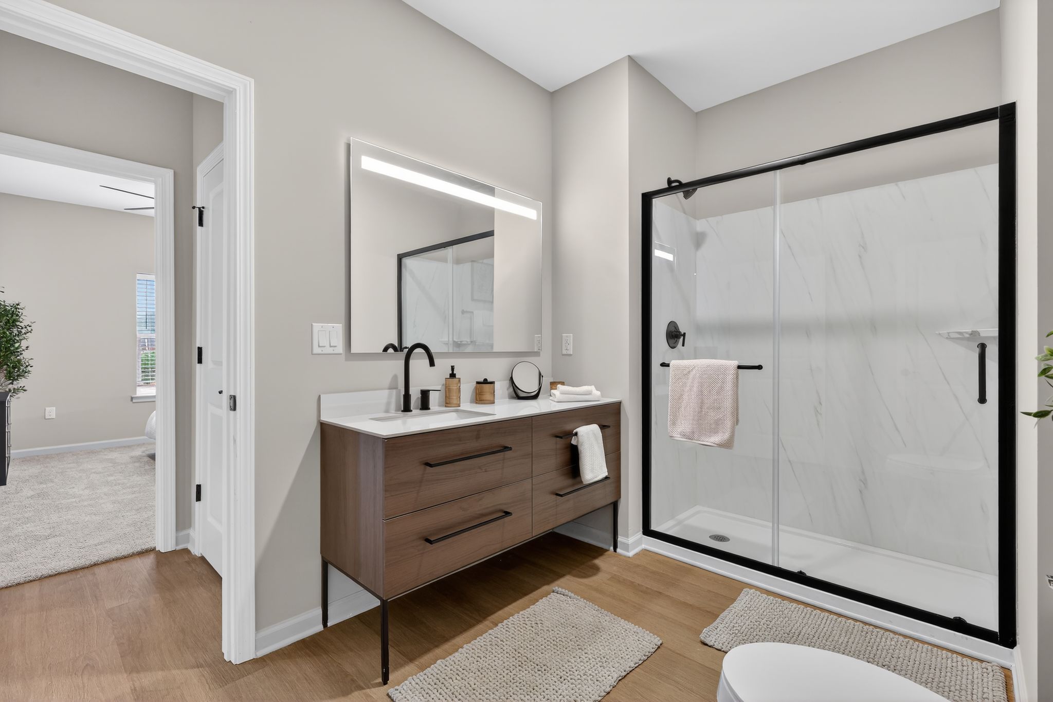 A modern bathroom featuring a sleek vanity with a large mirror and contemporary lighting. The shower area has a glass door, and there are neutral-toned walls and hardwood floors. A towel hangs on the vanity, and a plant adds a touch of greenery to the serene space.