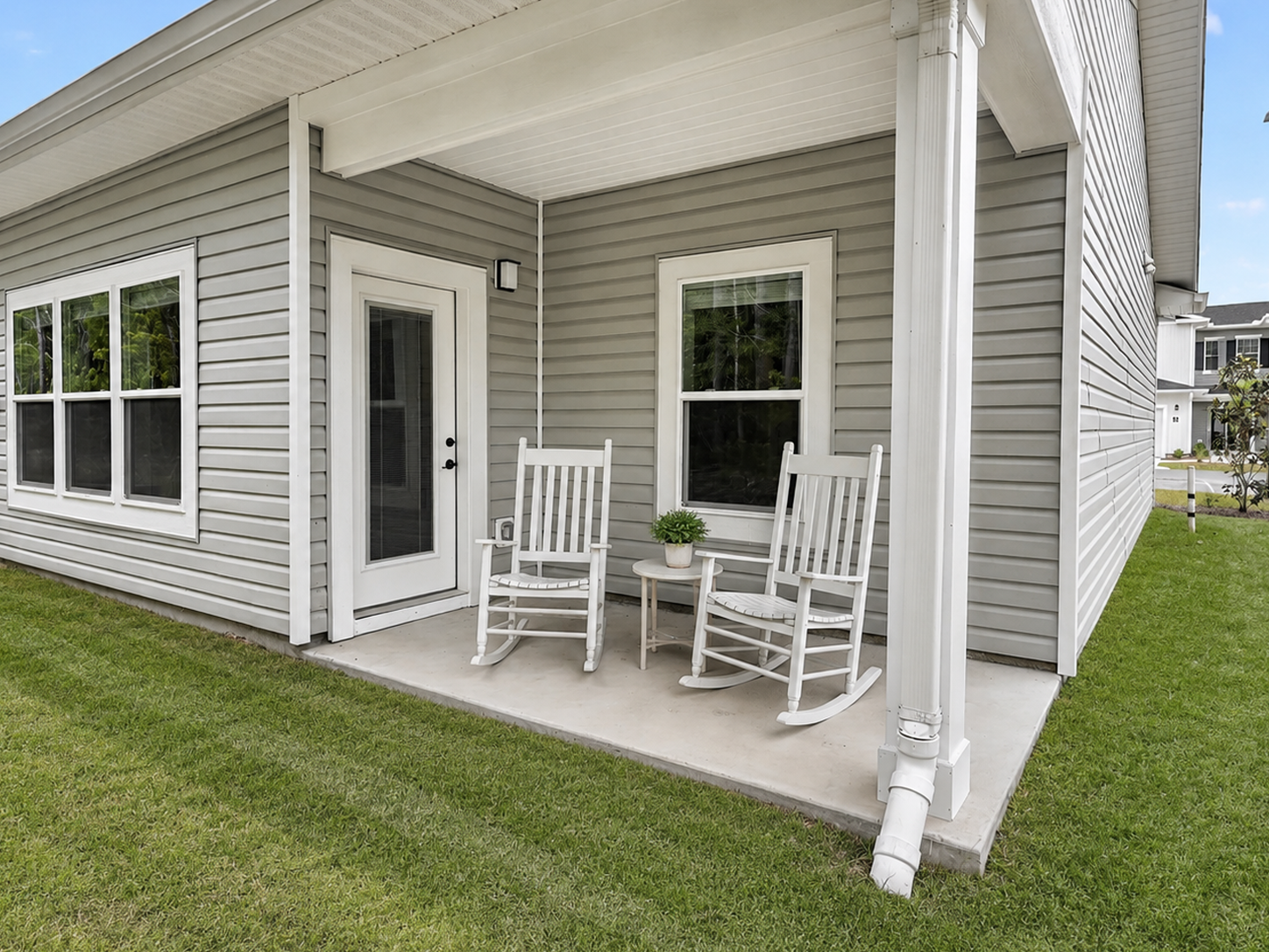 A cozy porch featuring two white rocking chairs positioned beside a small table with a potted plant. The porch is part of a modern gray home with white trim and large windows, situated on a grassy lawn. The scene conveys a welcoming and relaxing outdoor space.